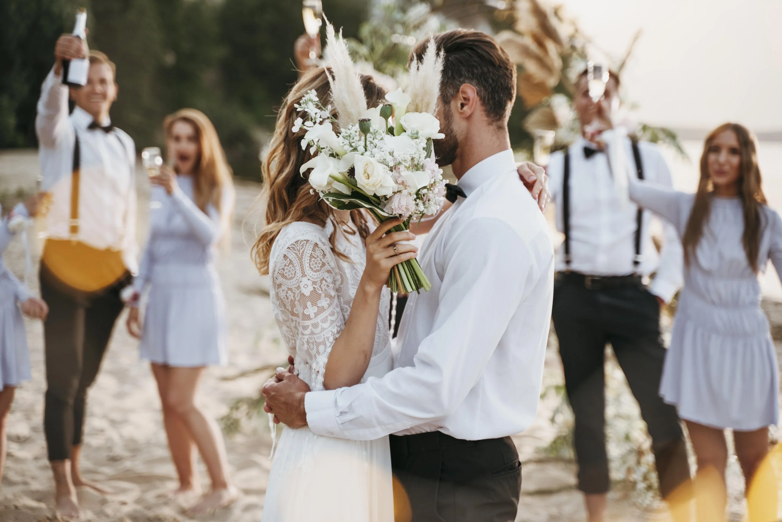 Bride and groom kissing with wedding party behind them on the beach