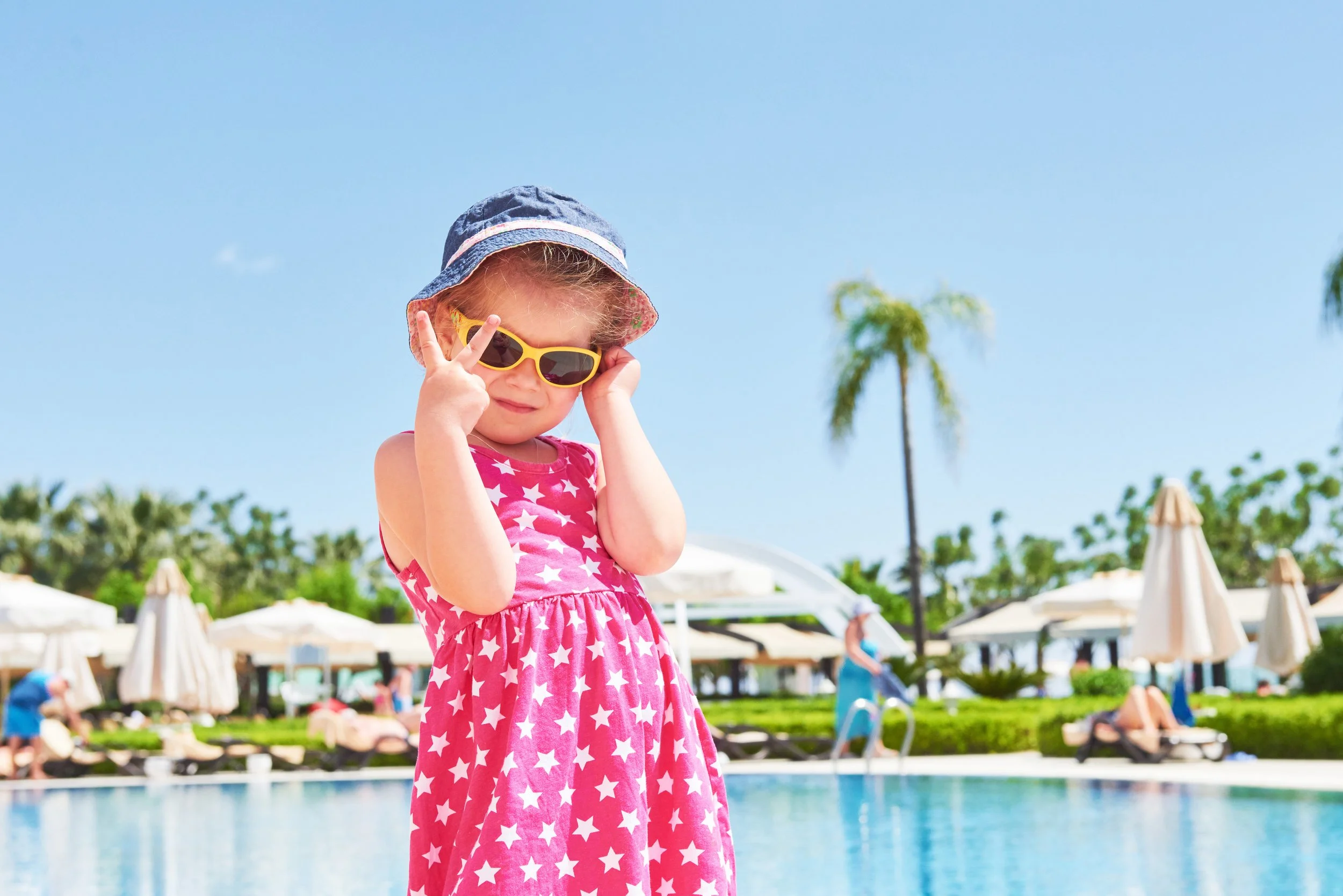 Little girl standing in front of a pool with sunglasses and hat