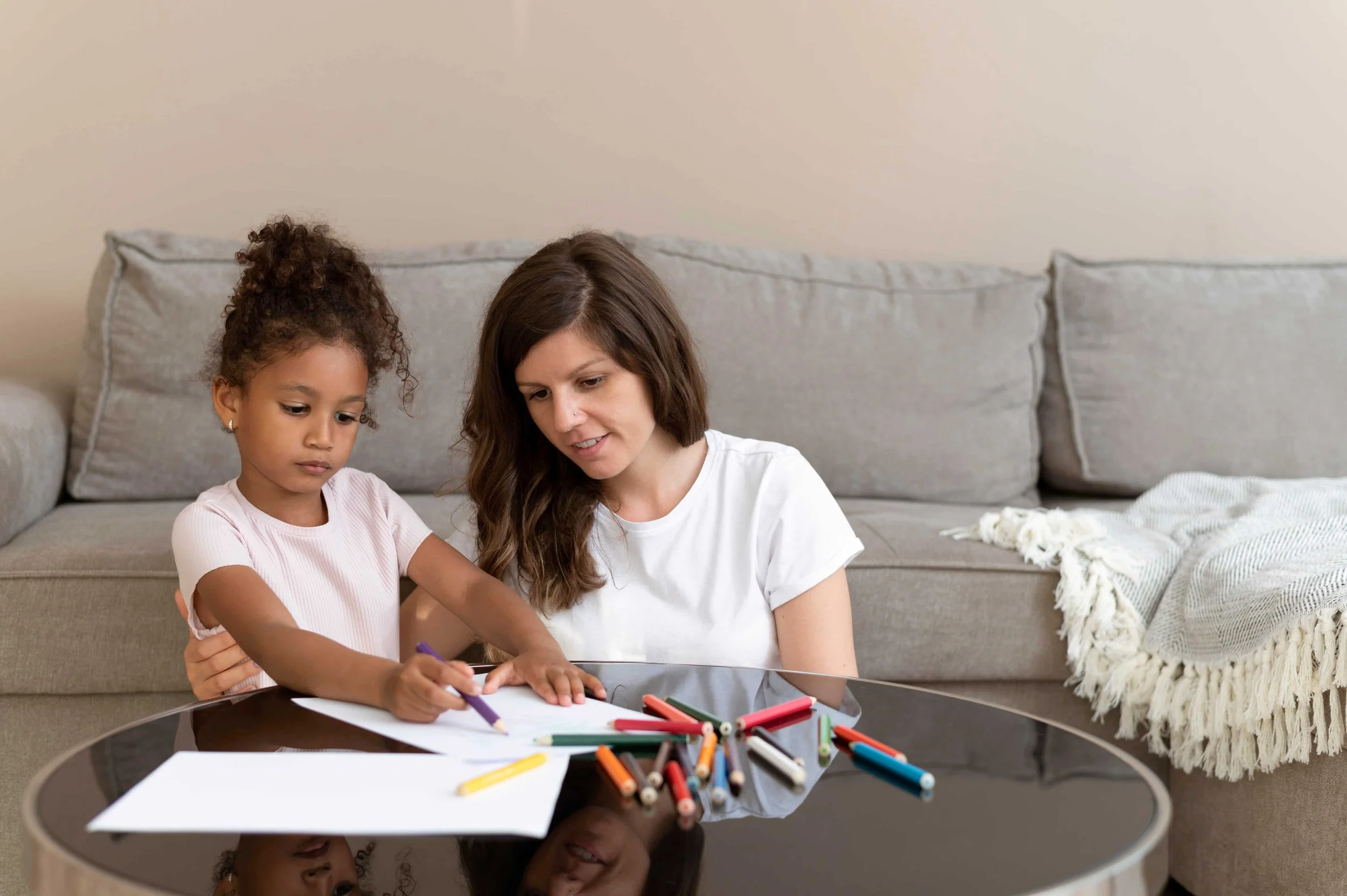 A woman and young girl drawing with markers on paper at a glass-top table in a living room.