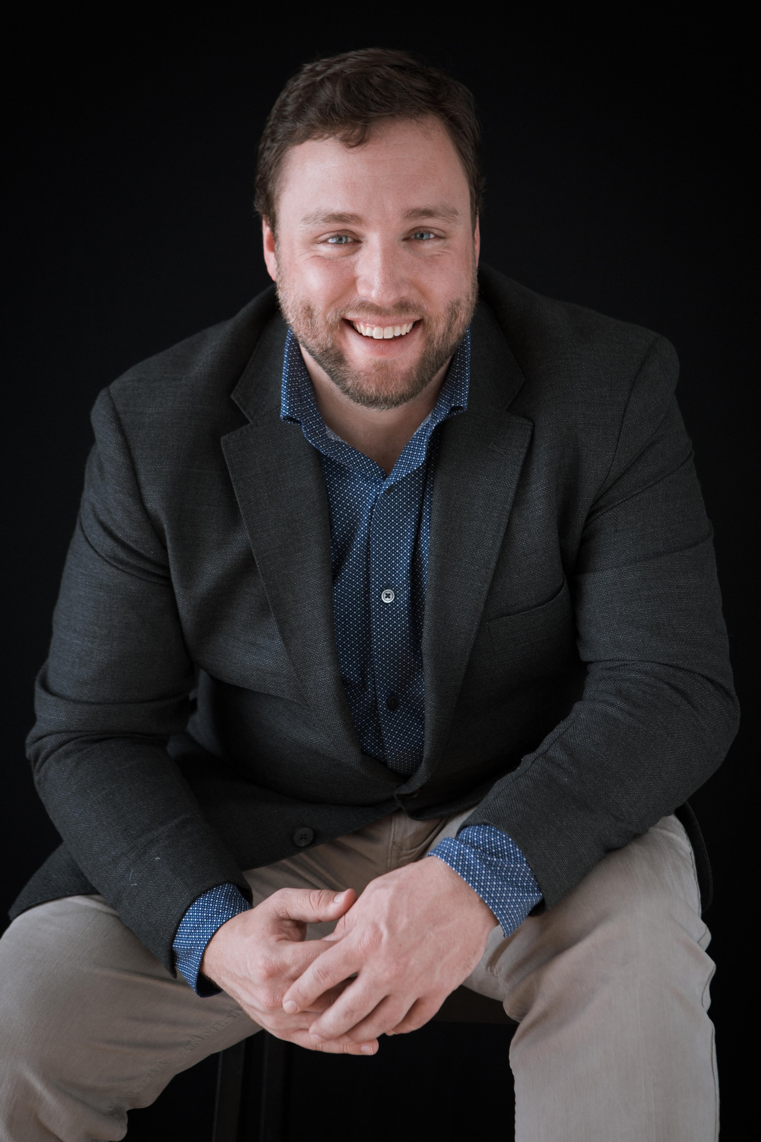 Portrait of a smiling man with a beard, wearing a dark blazer and light-colored pants, seated against a black background.
