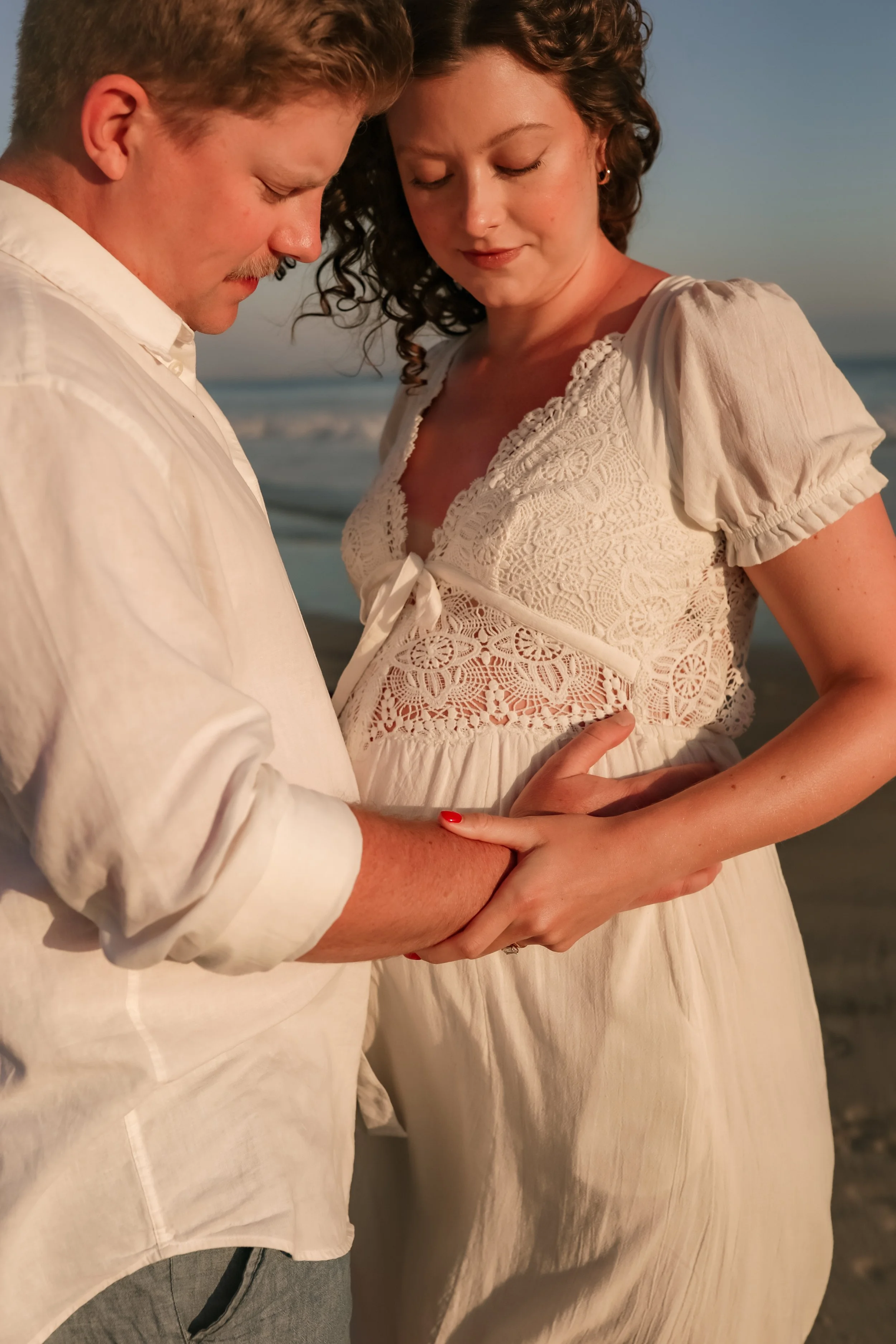 A man and woman sharing an intimate moment on the beach during sunset, with their foreheads touching and holding hands.