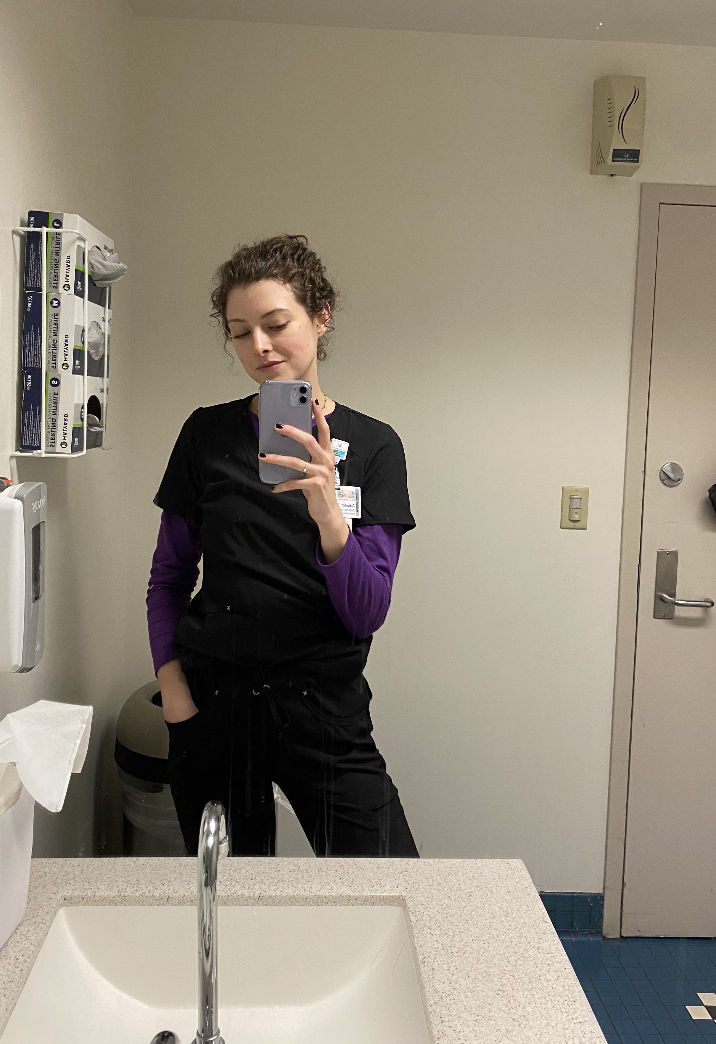 A young woman in a medical setting taking a selfie in front of a mirror. She is wearing black scrubs with a purple long sleeve shirt underneath and has a name badge on her chest. The background includes a soap dispenser, a paper towel dispenser, a trash bin, and a door.