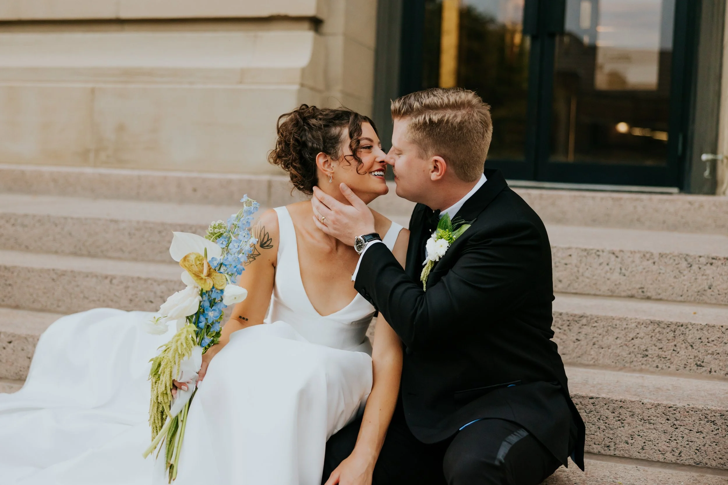 A bride and groom sitting on steps, touching noses and smiling, with the bride holding a bouquet of flowers.