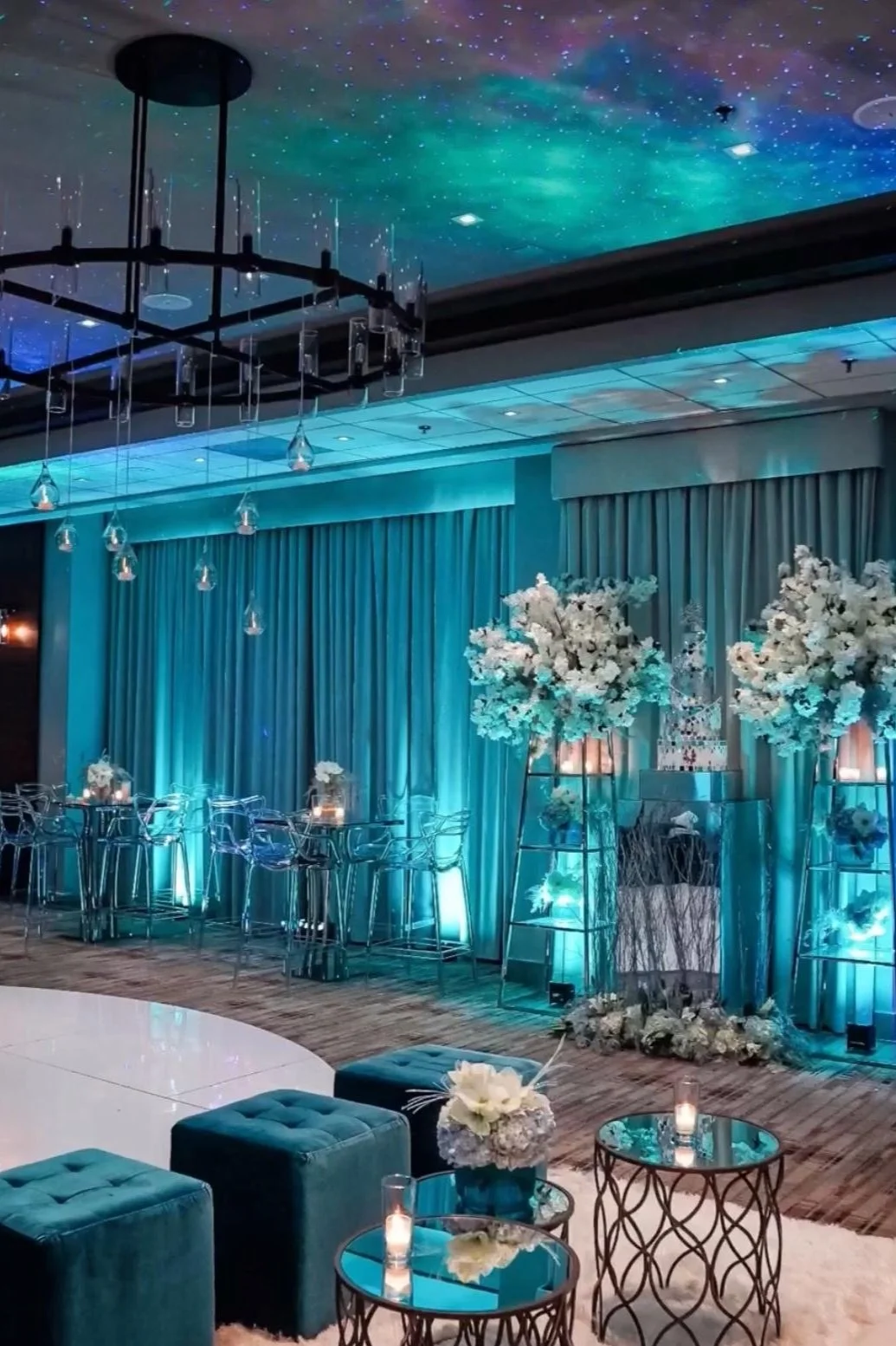 Elegant indoor event space with teal lighting, mirrored tables, blue upholstered seating, and floral decorations, featuring hanging glass lights and a starry ceiling.