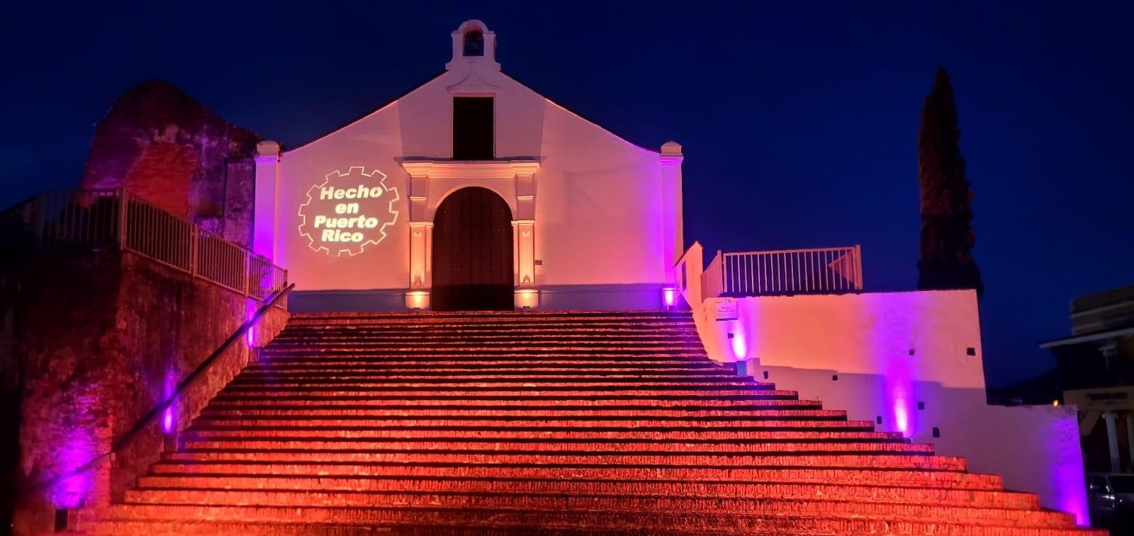 Illuminated steps leading to a small chapel with "Hecho en Puerto Rico" projected on the facade, lit by purple and amber lights, during nighttime.