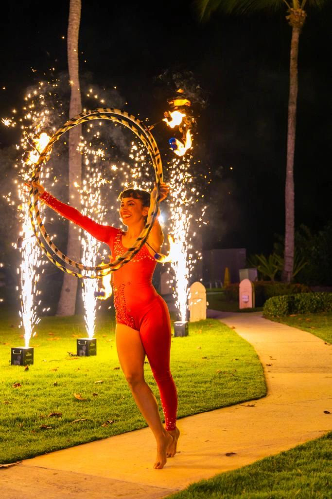 Performer in red costume spinning fire hoops with fireworks display in the background at an outdoor nighttime event.