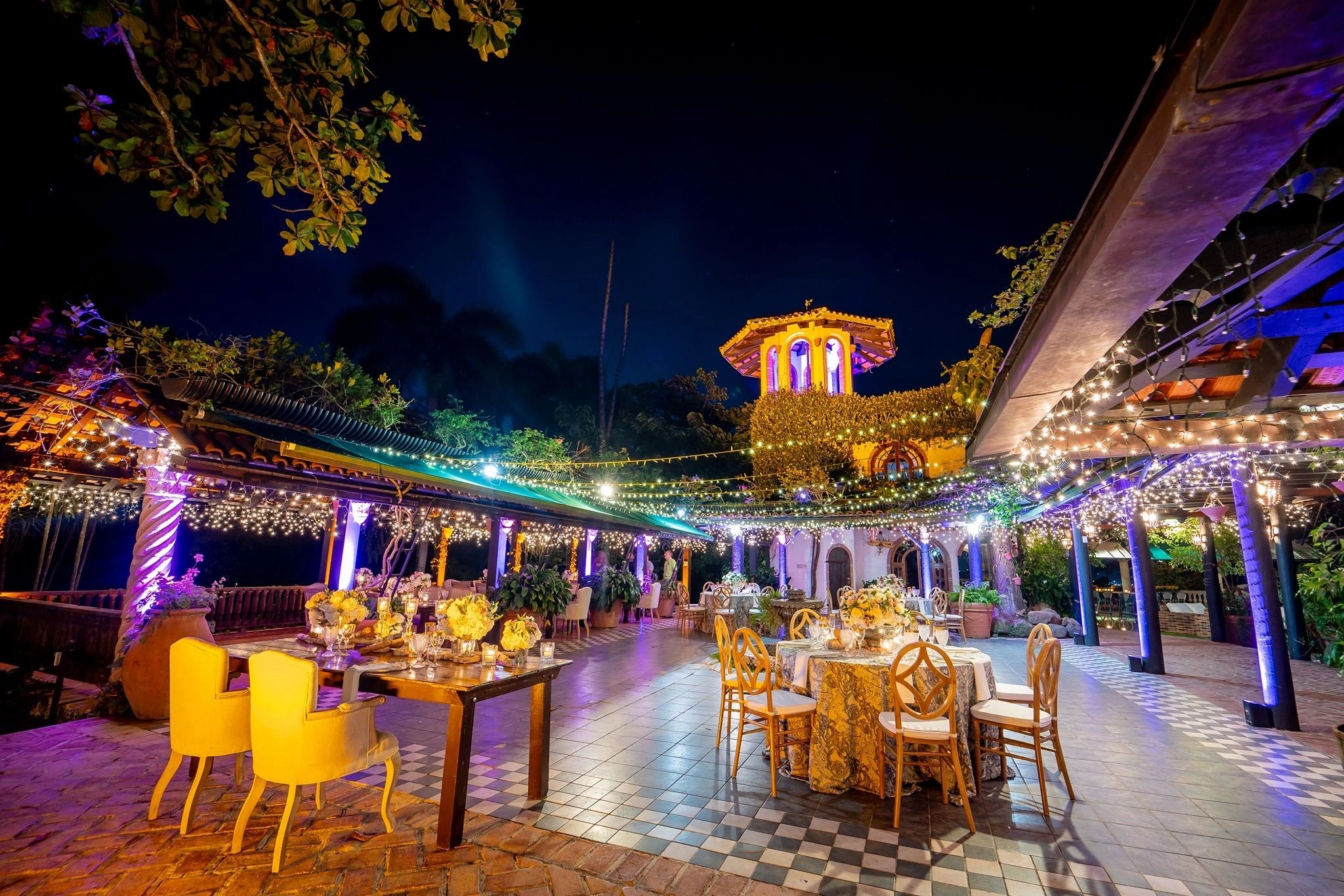 Outdoor wedding reception setup with elegant tables, chairs, and string lights, surrounded by lush greenery and a decorative tower in the background.