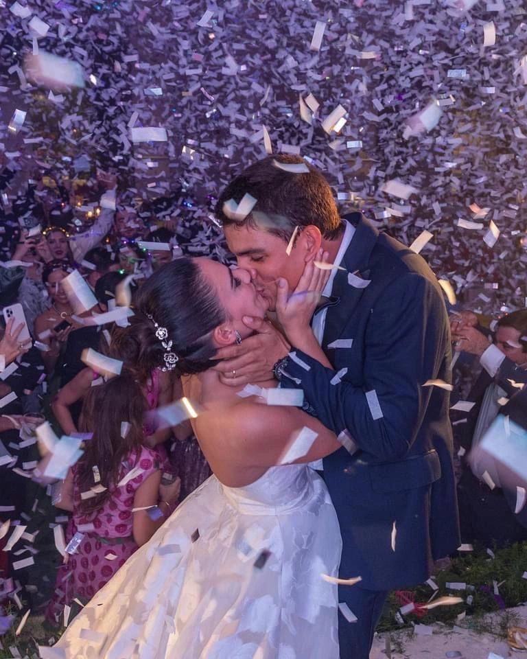 A couple in wedding attire kissing while confetti falls around them, surrounded by a crowd of celebrating people.