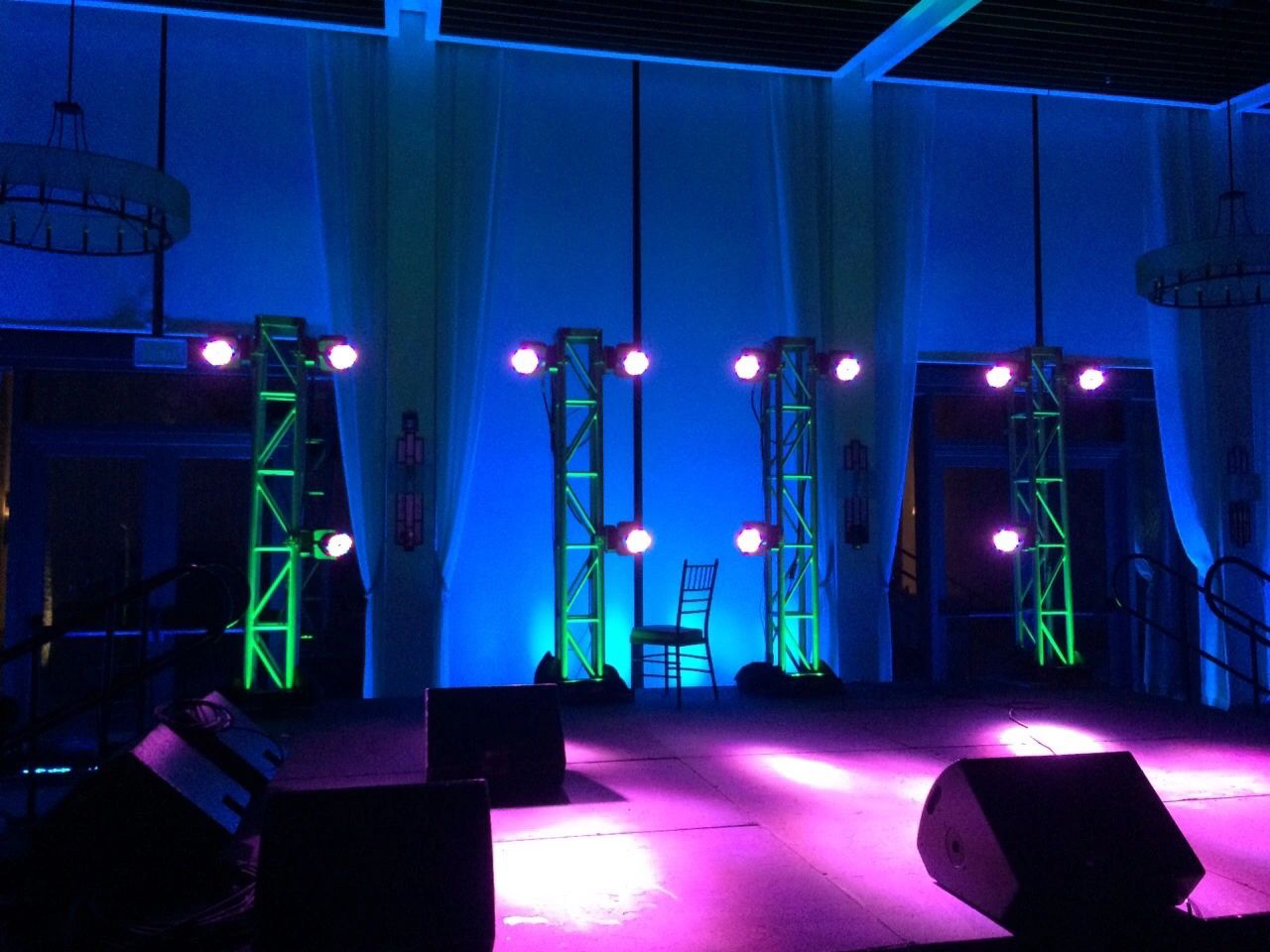 Stage setup with purple lighting, green trusses, and a single chair. There are two large speakers and a backdrop with curtains.