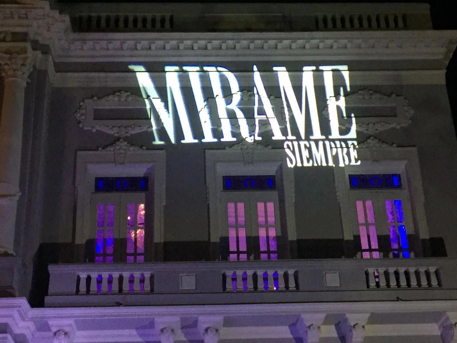 Illuminated facade with the words "MIRAME SIEMPRE" projected on a building, featuring ornate architectural details and purple-lit windows.