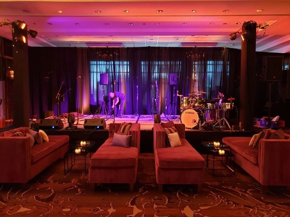 Cozy lounge with dim lighting and a stage setup for live music, featuring a drum set, microphones, and sofas.