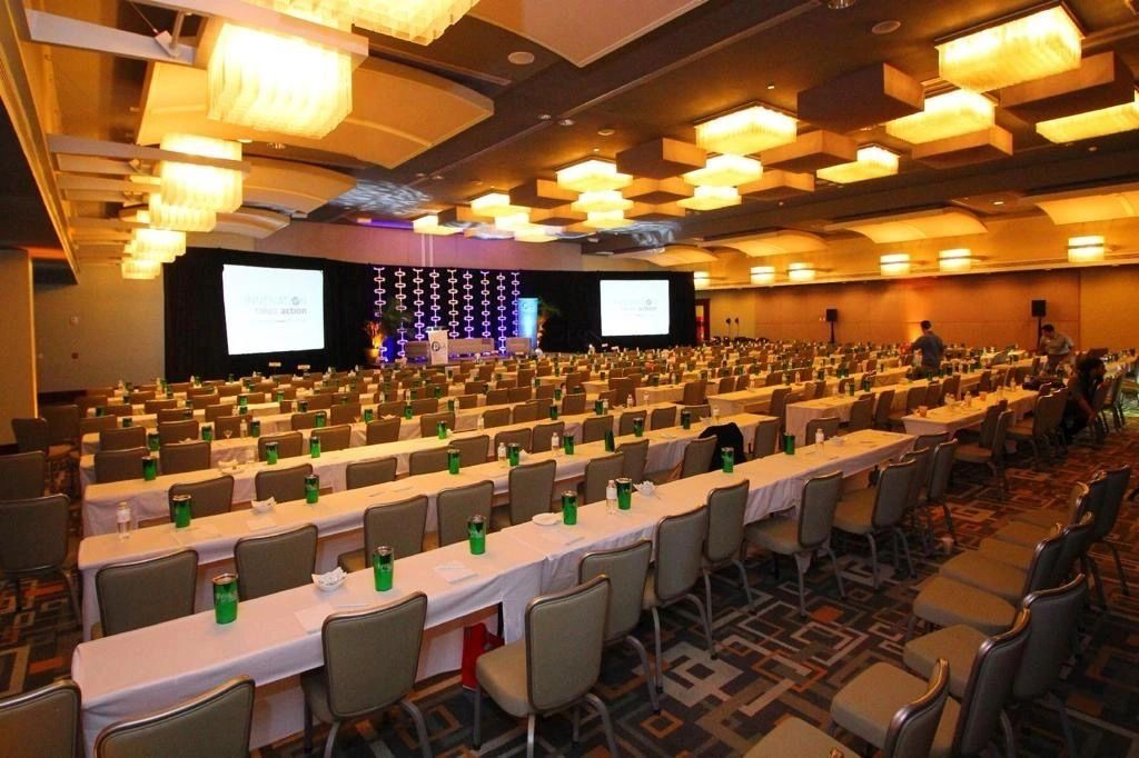 Large conference room with rows of tables and chairs, projectors on stage, and ceiling lights.