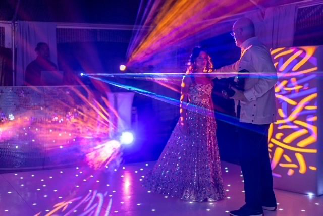 A dimly lit event space featuring a woman in a sparkly dress and a man interacting, surrounded by colorful laser lights and decorative patterns on the walls.