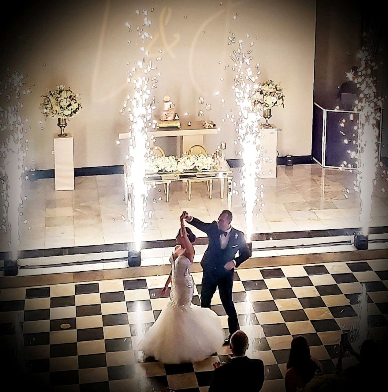 Bride and groom dancing under fireworks indoors at their wedding reception on a checkered floor.