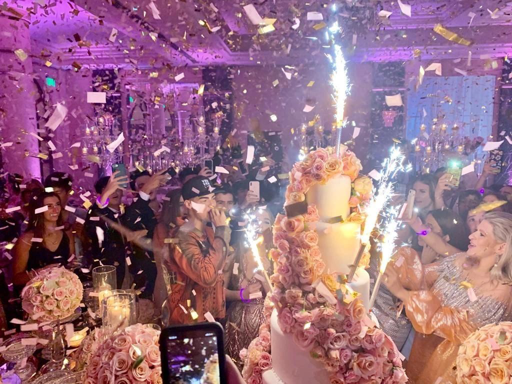 Celebration scene with people, a large floral cake with sparklers, and confetti falling.