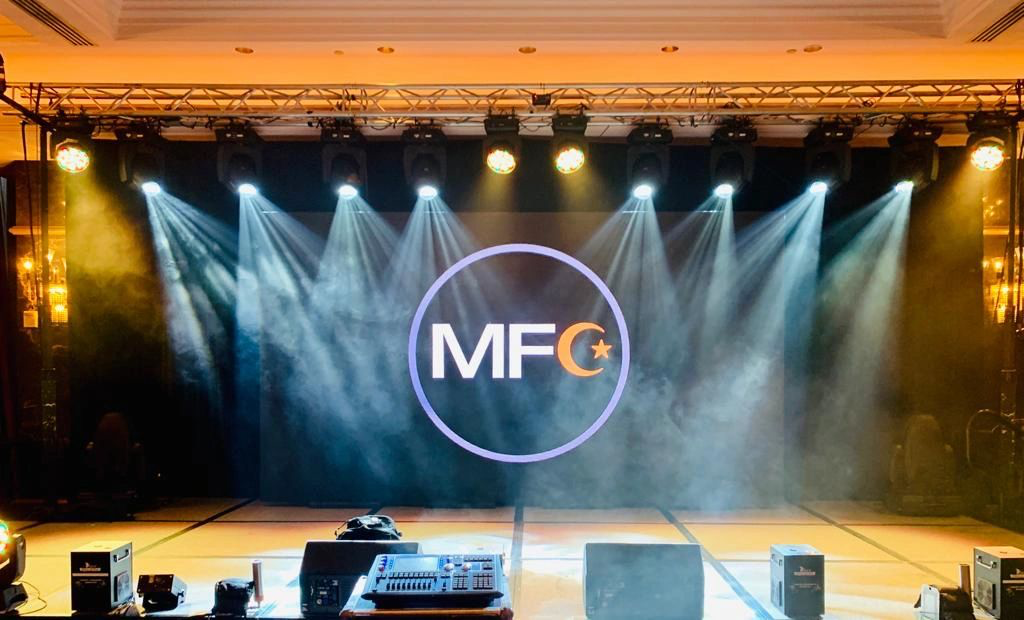 Stage setup with "MFC" logo on LED screen, surrounded by spotlights and sound equipment.