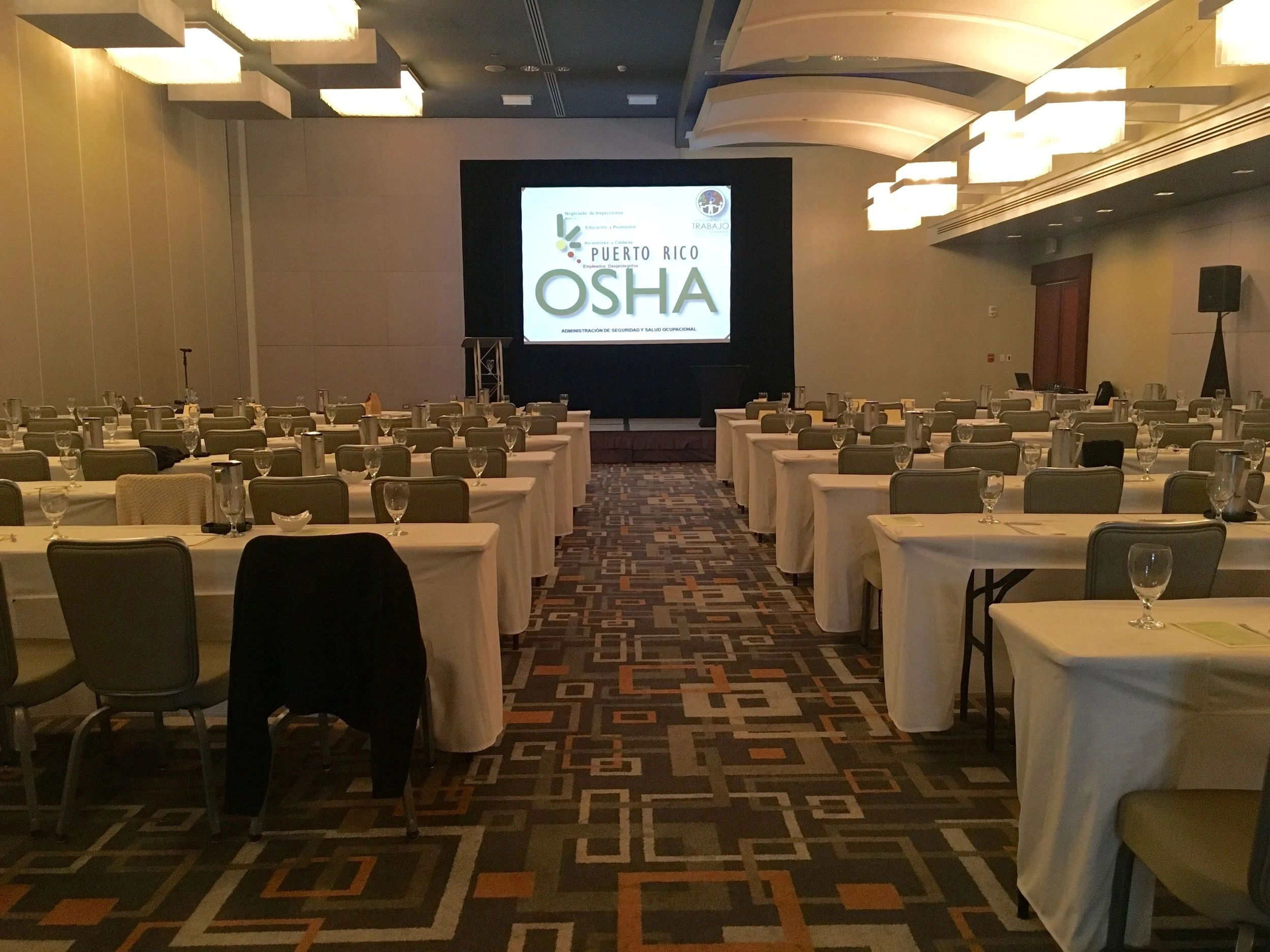 Conference room with rows of tables and chairs, set up for a Puerto Rico OSHA event. Tables are covered with white cloths and equipped with water glasses and notepads. A presentation screen displays the event logo.