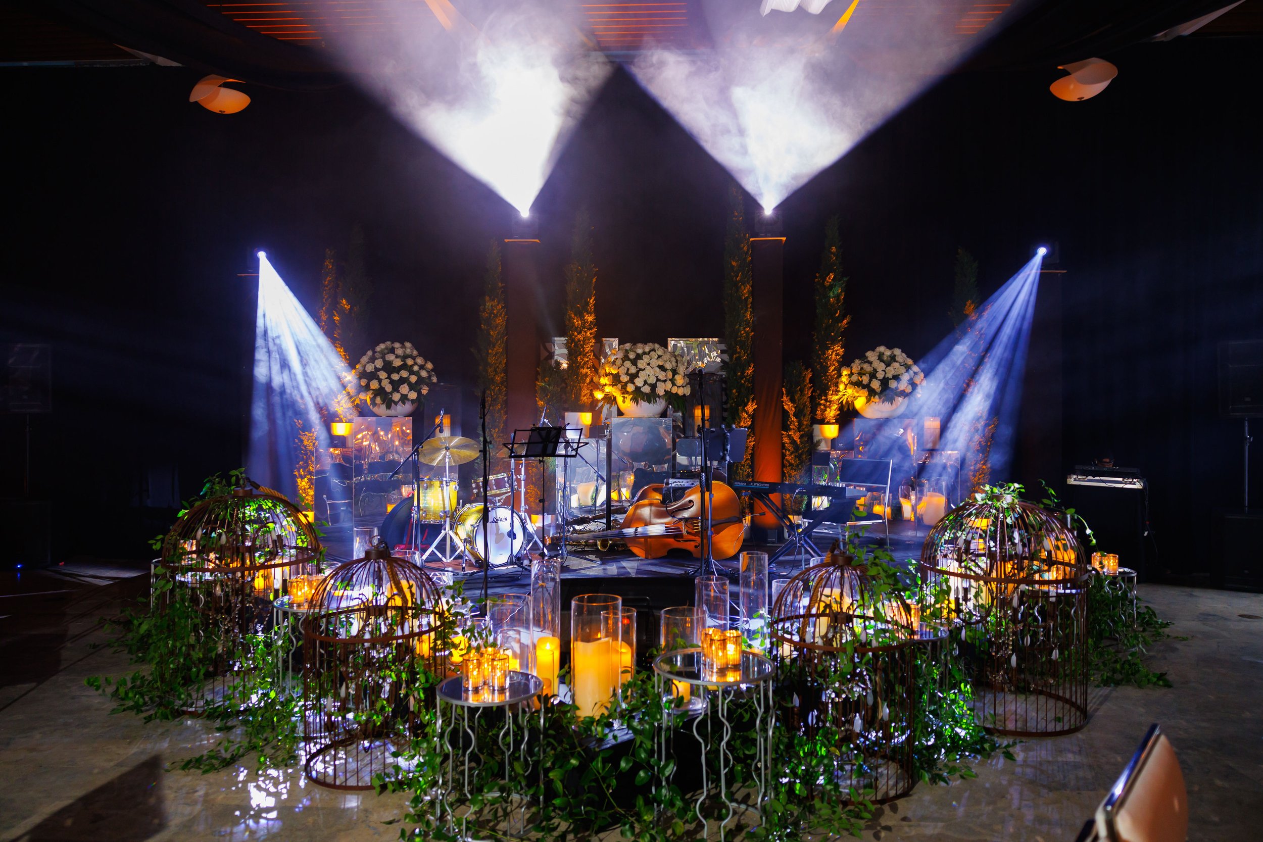 Our triangle shaped stage decorated with an arrangement of flowers, leaves, and candles. There are stage lights and ambient lights illuminating the stage with a radiant glow. On the stage there are musical instruments.