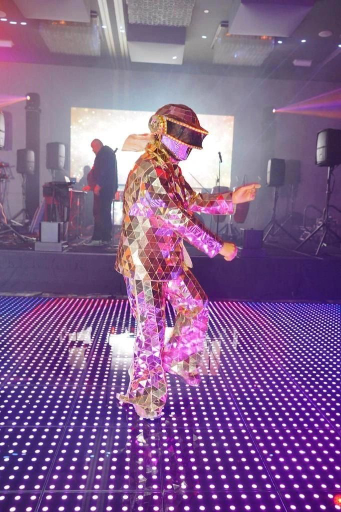Person in a reflective, geometric costume dancing on an illuminated floor with disco lights, surrounded by sound equipment and a DJ in the background.