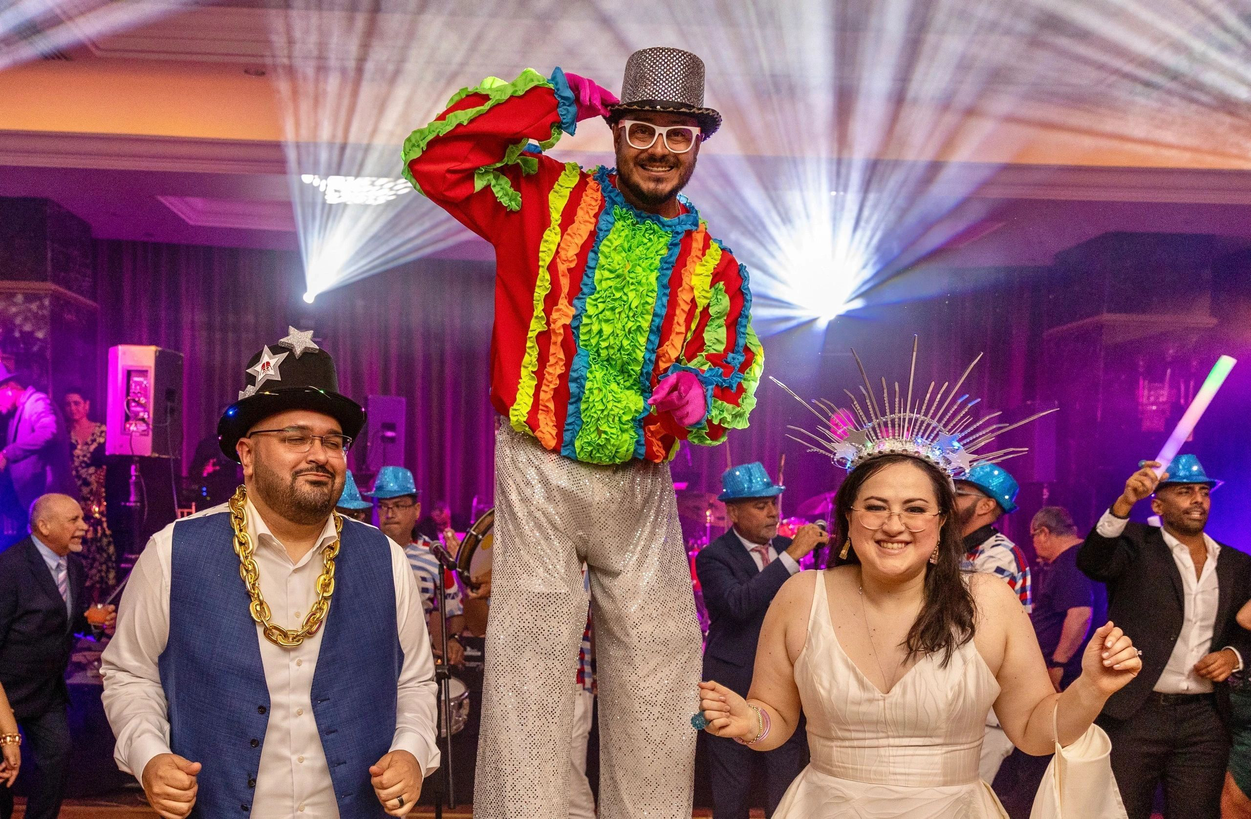 A festive celebration with a performer in a colorful costume and silver sequin pants, surrounded by people in costumes and hats, under bright stage lighting.
