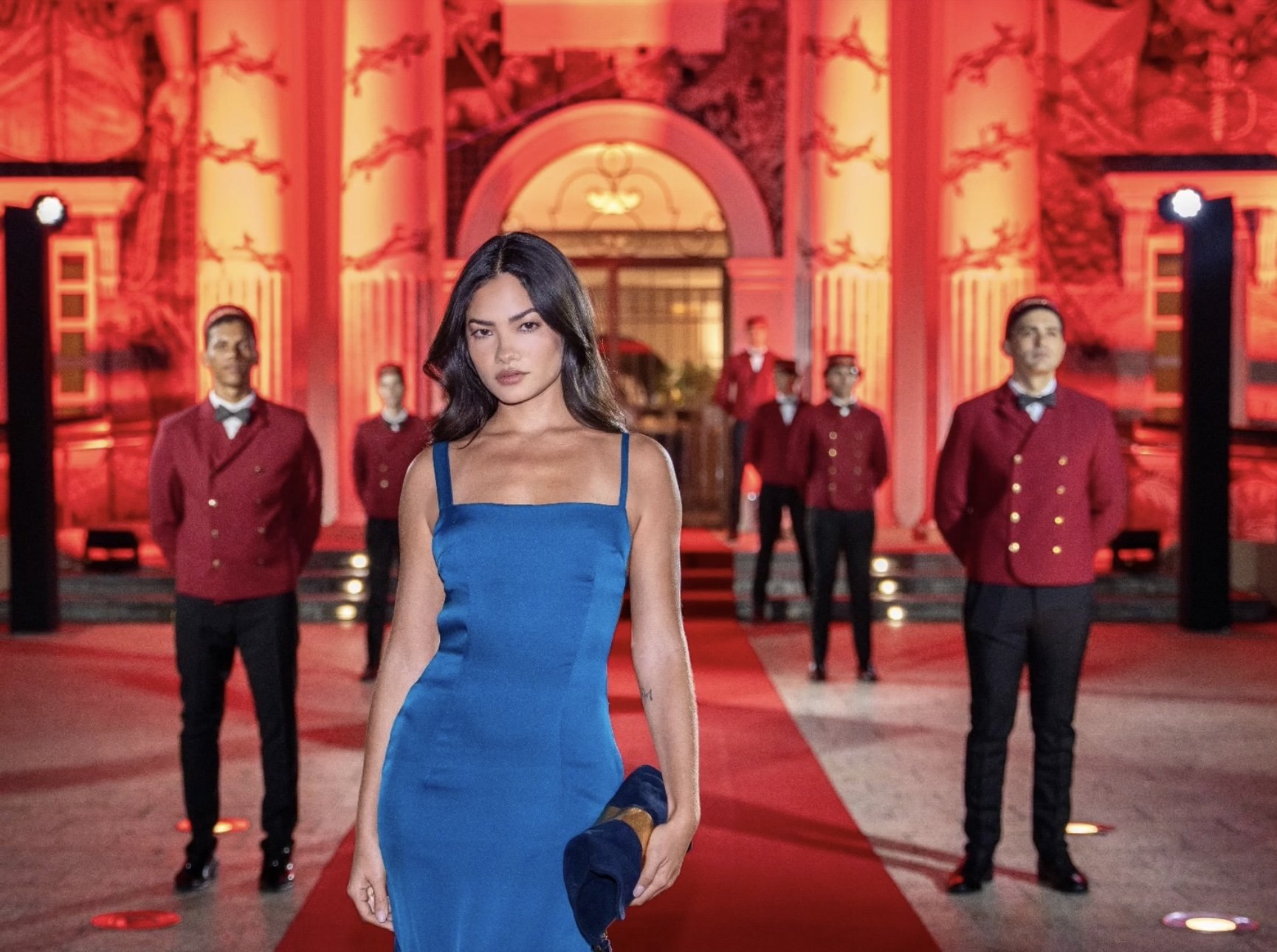 Model with blue dress standing in front of five men with red Cartier butler jackets with red ambient architectural lighting