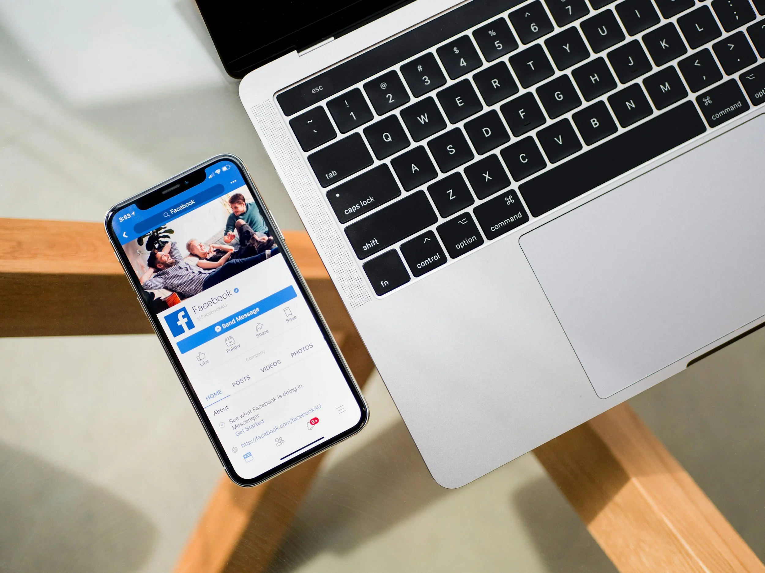 A laptop and a smartphone on a wooden desk, displaying Facebook's homepage, with the phone showing a group of young people sitting together.