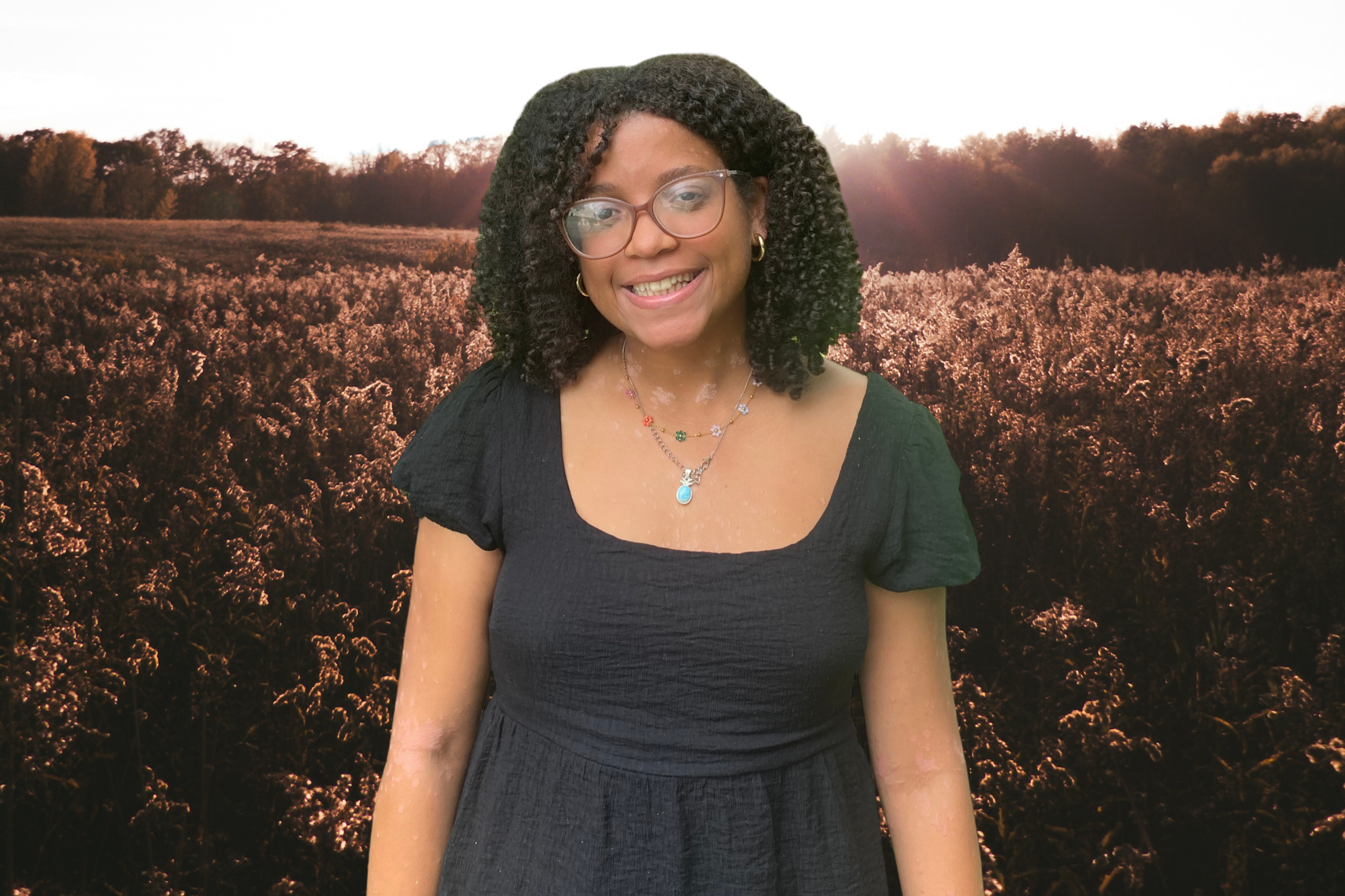 A woman with glasses and curly black hair smiling, standing outdoors during sunset with a field of trees in the background.