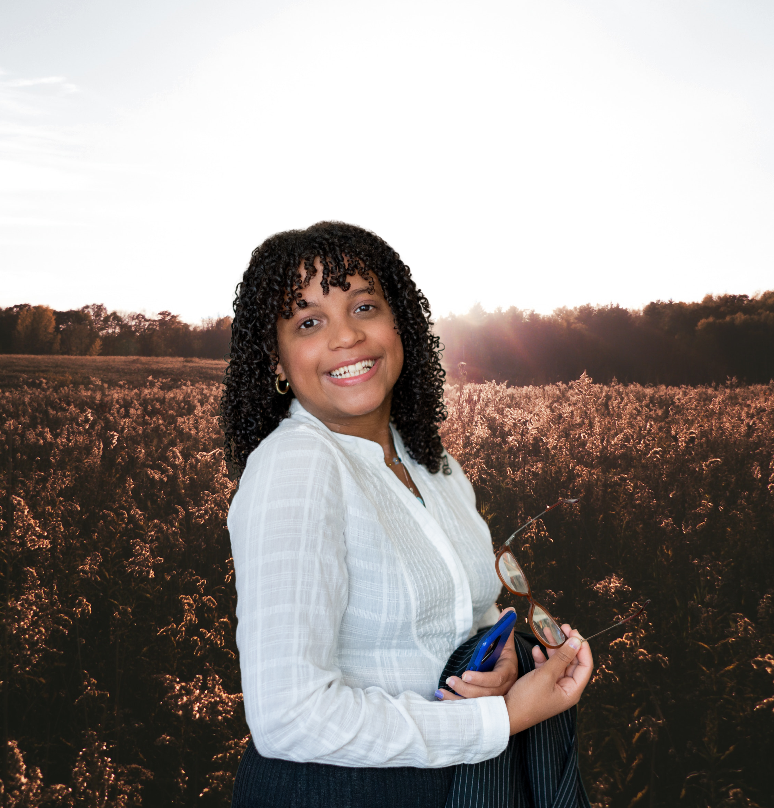 Close-up of a smiling woman with curly hair wearing a white shirt, holding glasses, a phone, and a notebook, with a scenic background of trees and a sunset sky.