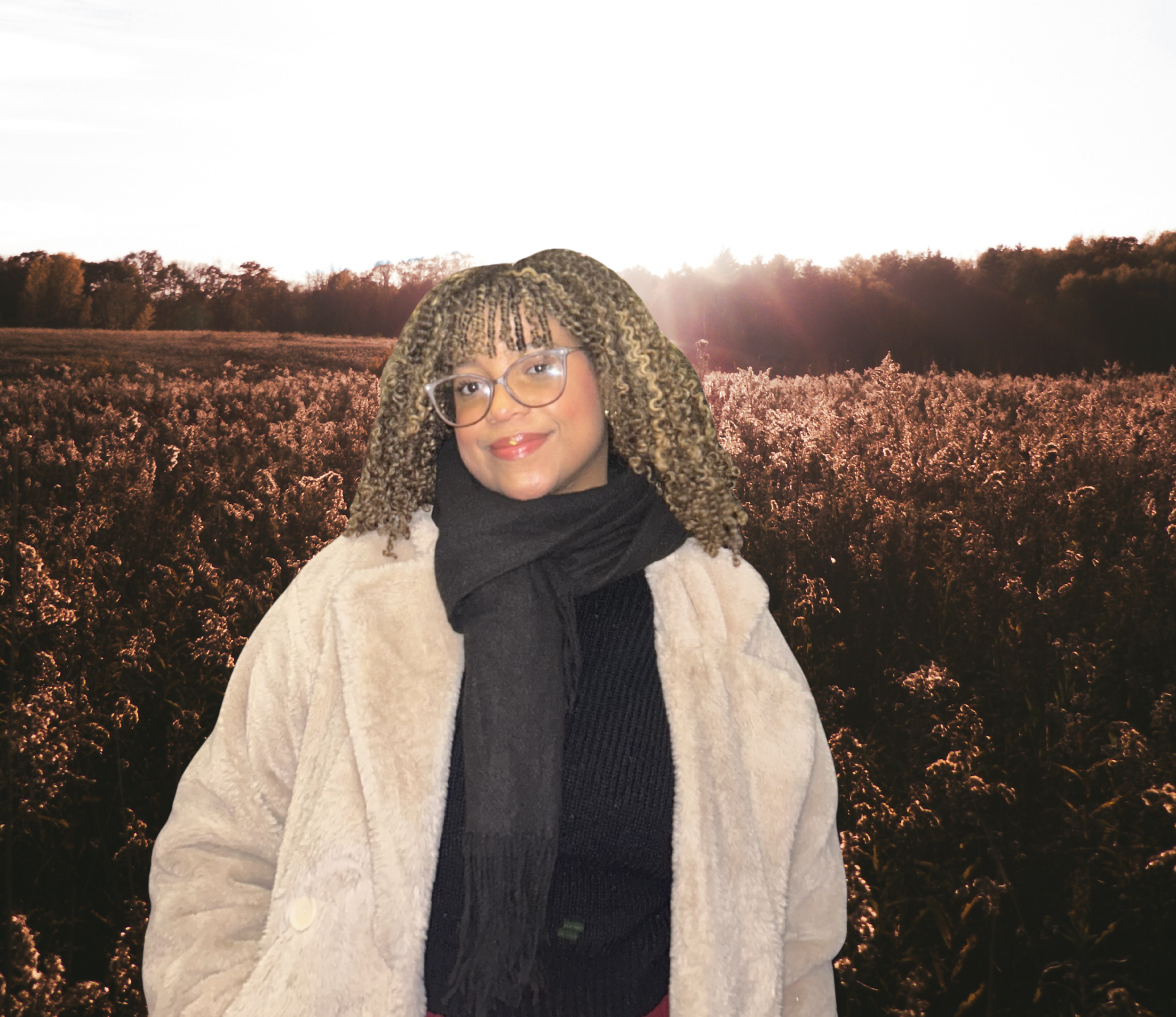 A woman with curly blonde hair and glasses standing outdoors at sunset or sunrise with trees and a field of flowers in the background.