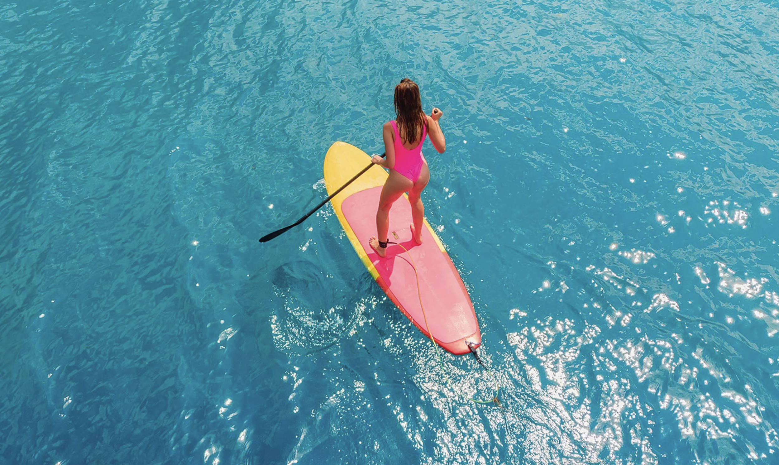 A woman in a pink swimsuit stands on a Stand Up Paddle Board in clear blue water at Coral Cove.
