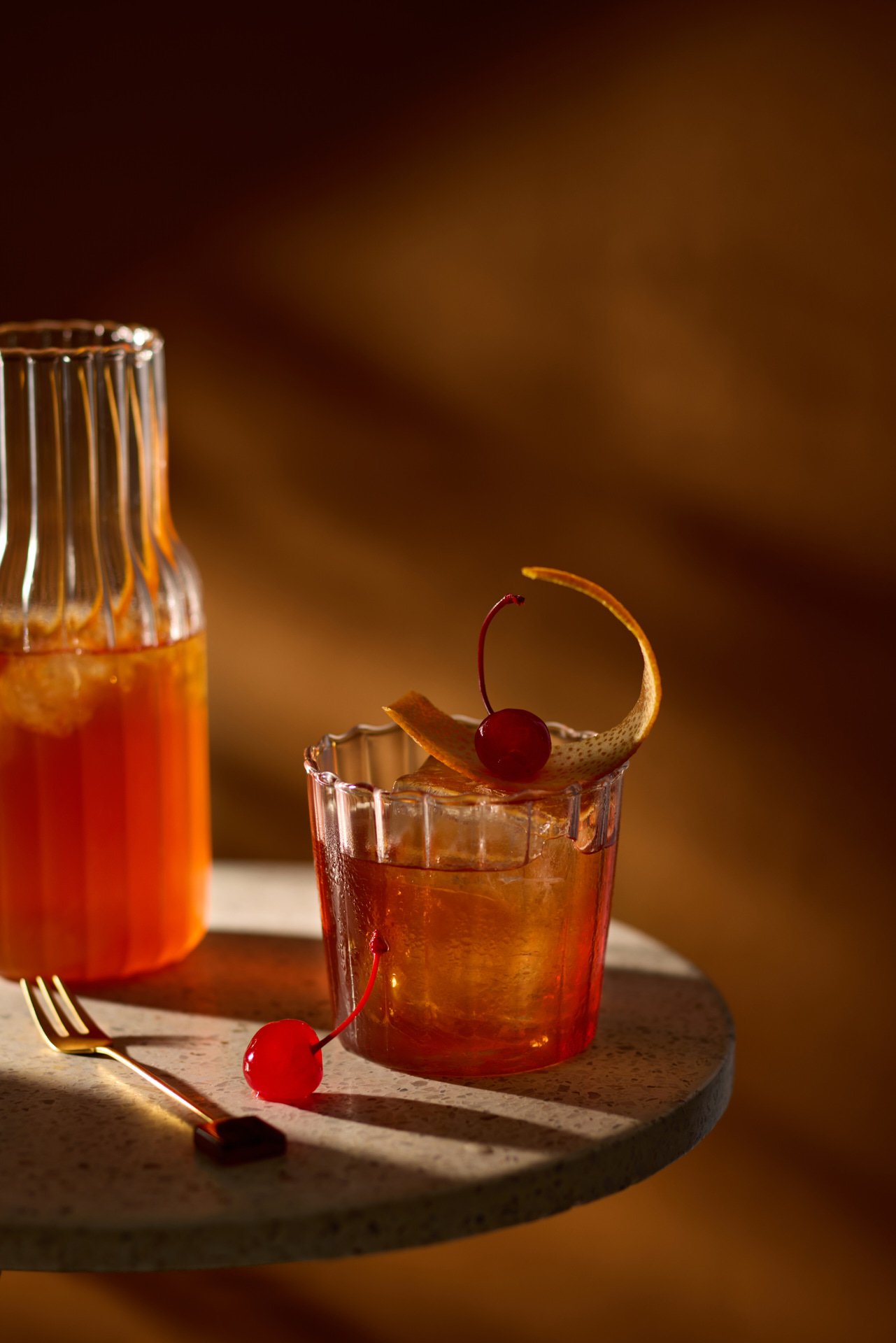 Whiskey cocktail with cherry and orange twist garnish, next to a carafe of orange beverage on a round table.