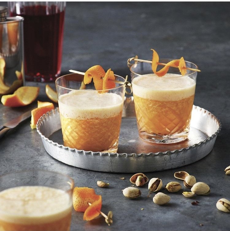 Two orange cocktails with foam, garnished with orange peel twists in glasses on a silver tray, surrounded by citrus peels and pistachios.