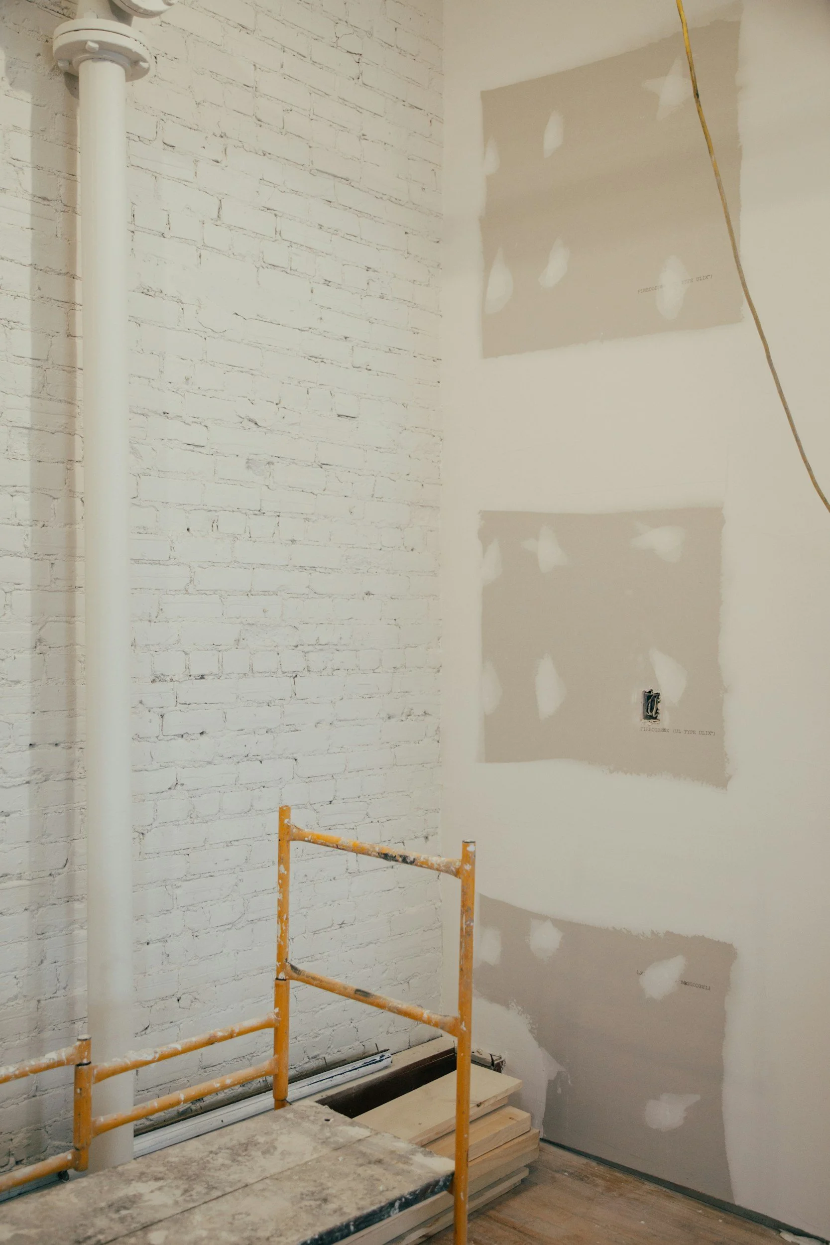 Interior wall under construction with drywall and exposed brick, a vertical white pipe, a yellow safety barrier, and wooden planks on the floor.