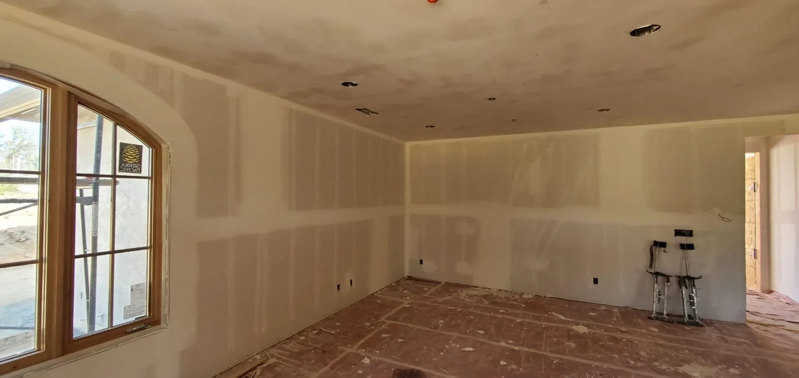 Interior of a room under construction with drywall installation, a window on the left, and electrical wiring visible on the wall to the right.