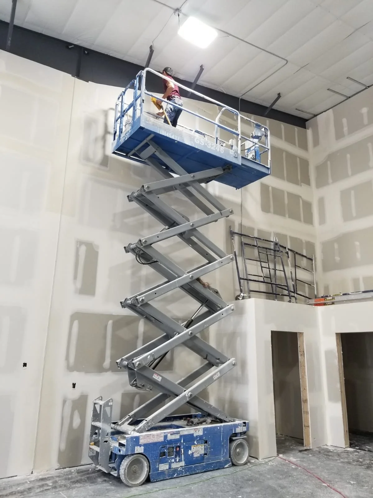 A worker is on a blue scissor lift working on an unfinished wall with drywall in a construction site.