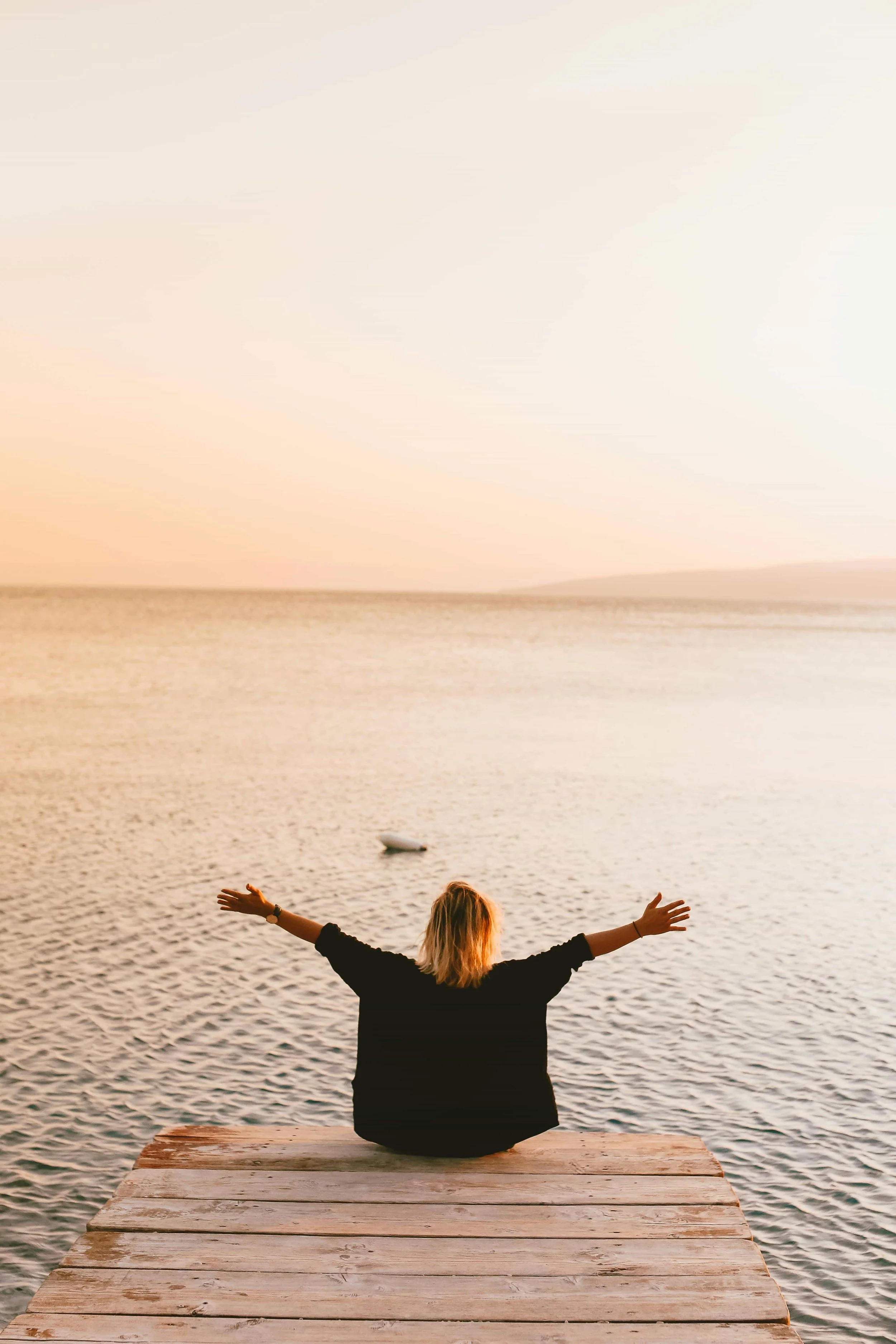 What It Means to Heal Trauma Through the Body -Woman sitting on a dock by a lake raising her hands