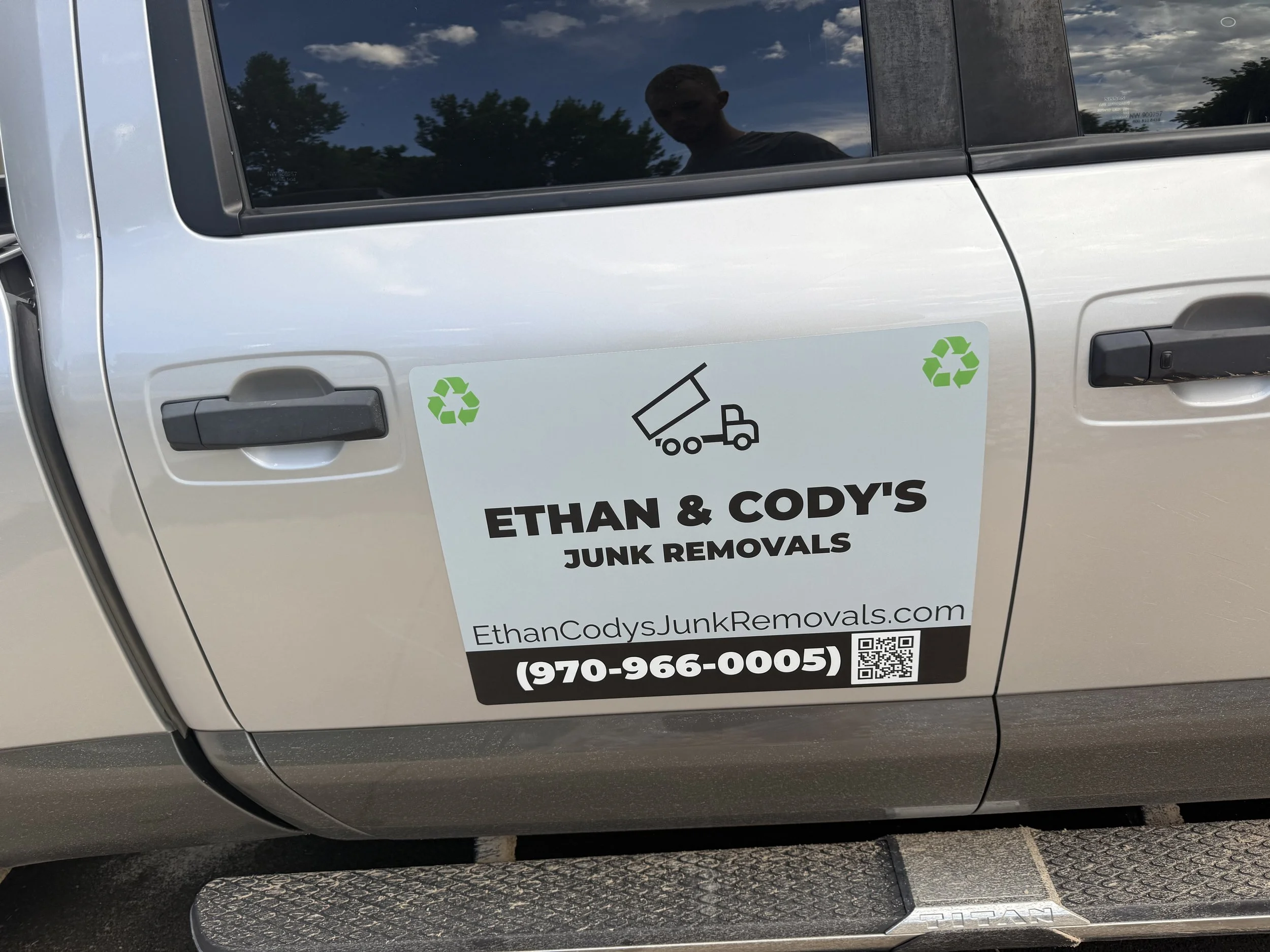 Silver vehicle with a bumper sticker advertising Ethan & Cody's Junk Removals, including company website, phone number, and recycling symbols.