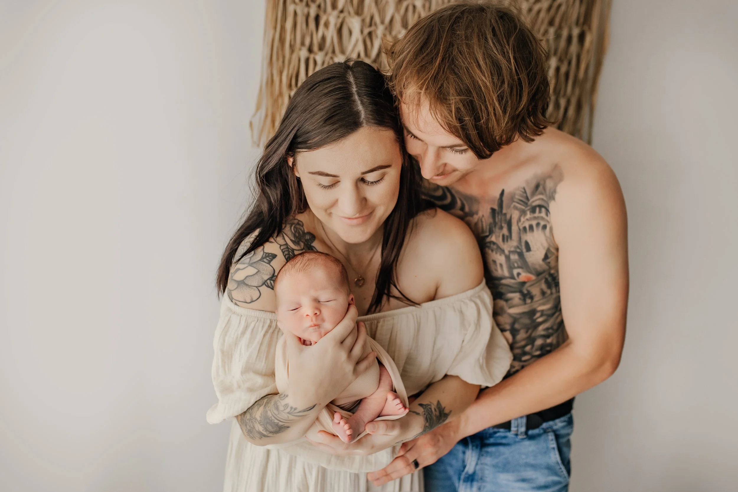 A woman holding a sleeping baby while a man embraces them; both have visible tattoos. They are indoors against a neutral backdrop.