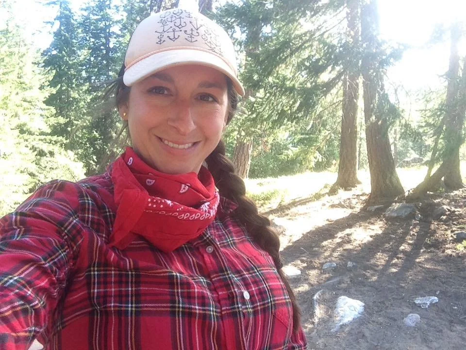 Marissa smiling outdoors in a forest, wearing a plaid shirt, a white cap with symbols, and a red bandana around her neck.