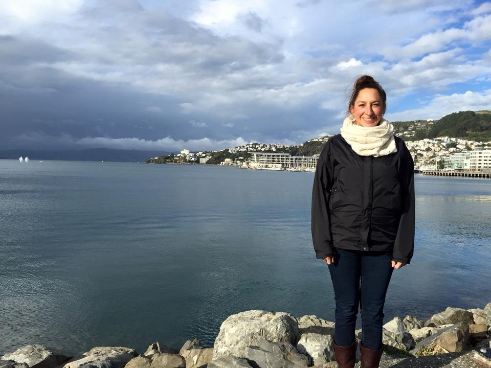 Marissa standing on rocks by a calm body of water, with a city and hills in the background under a partly cloudy sky.