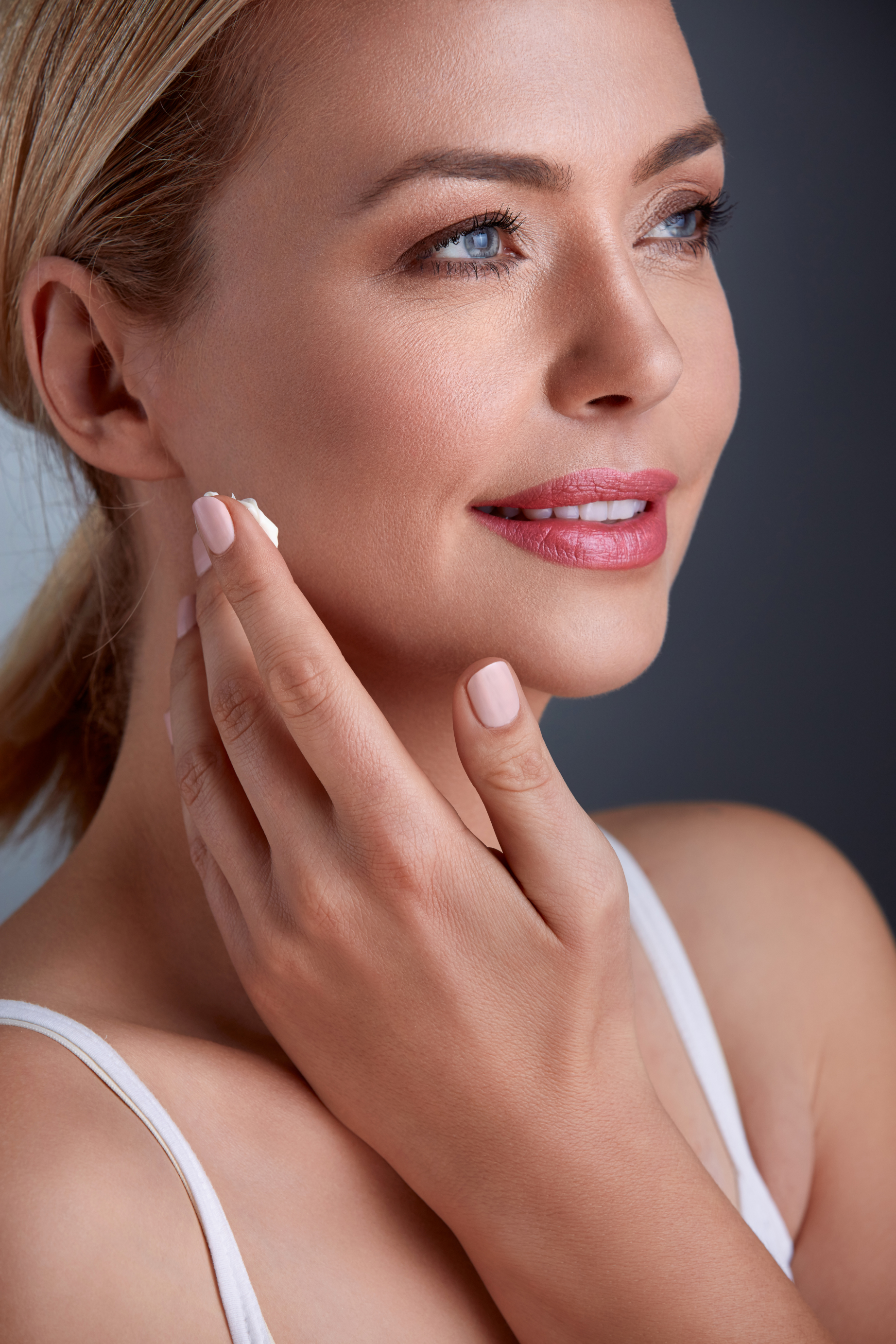 Close-up of a woman with blonde hair, applying lotion to her face, wearing a white tank top, with a dark background.
