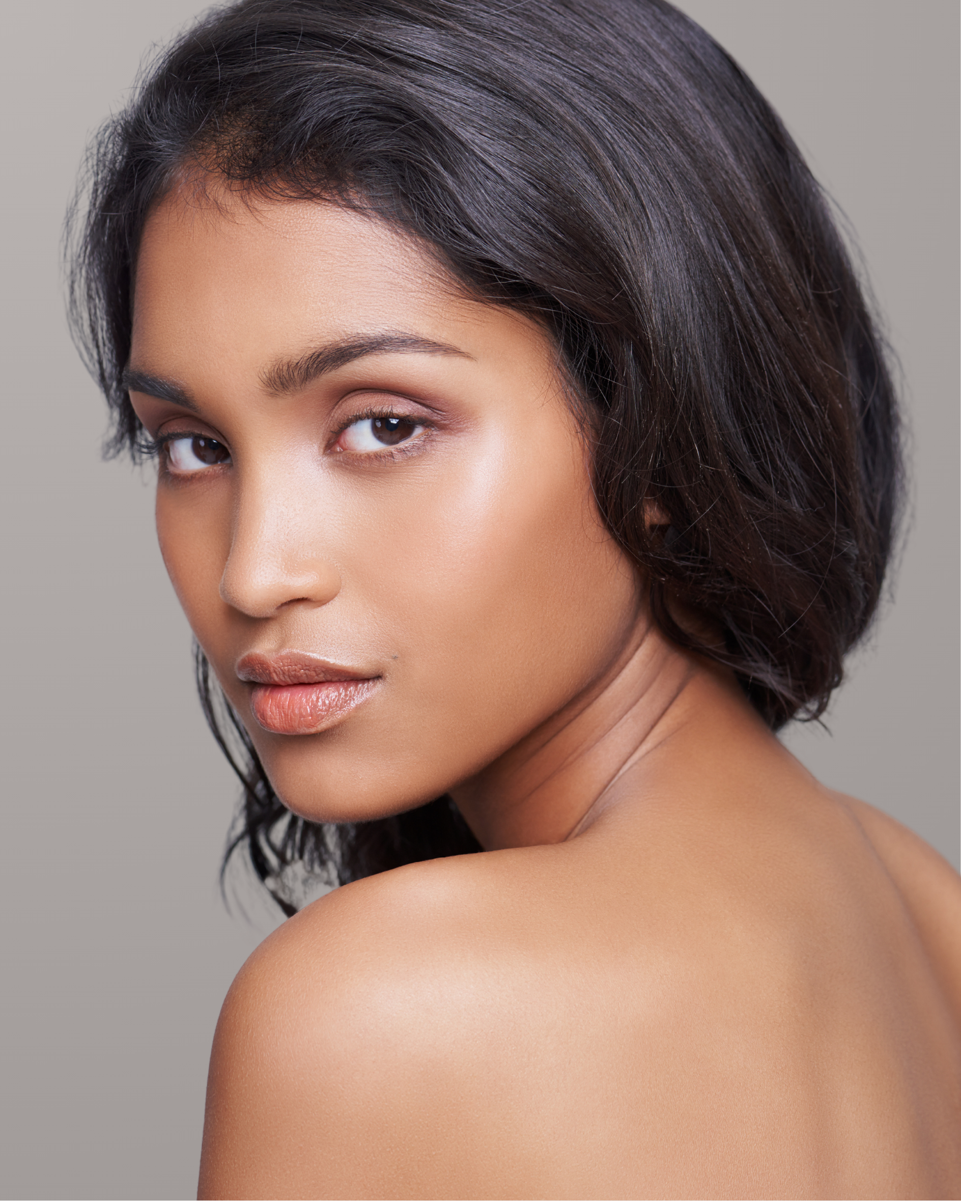 Close-up portrait of a woman with medium skin tone, dark brown hair, and subtle makeup, looking over her shoulder with a neutral expression.