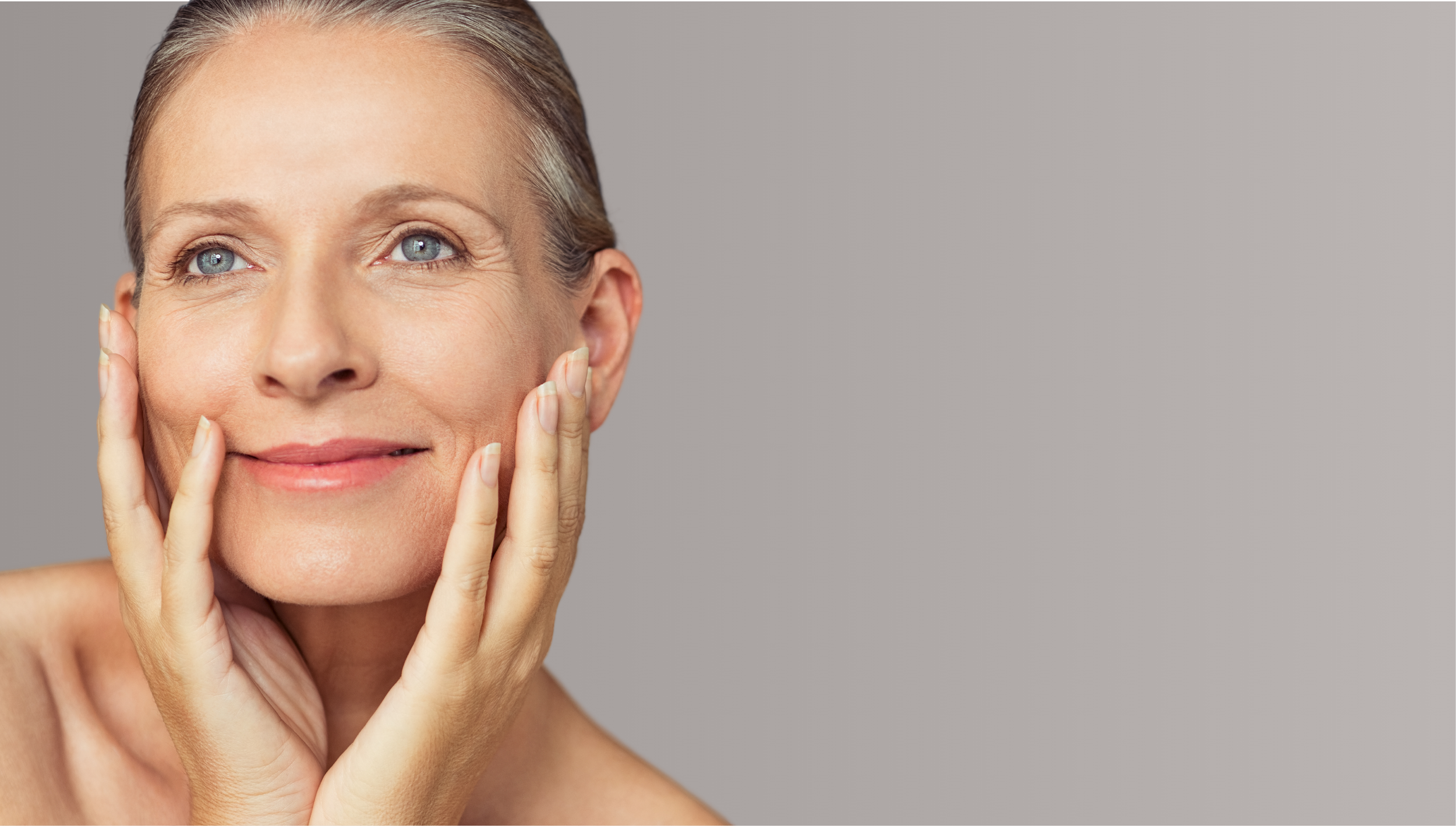Close-up of a middle-aged woman with clear skin, blue eyes, and short blonde hair, touching her face with both hands, smiling softly against a neutral gray background.