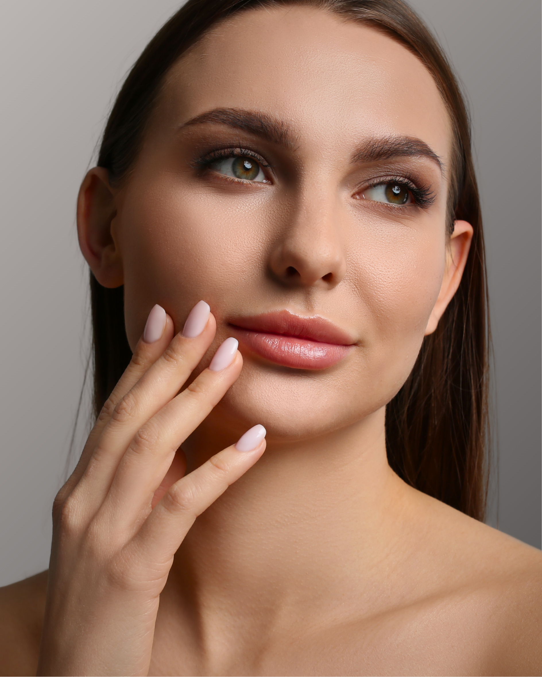 Close-up of a woman with smooth skin, brown hair, and makeup, touching her face gently with her fingers.
