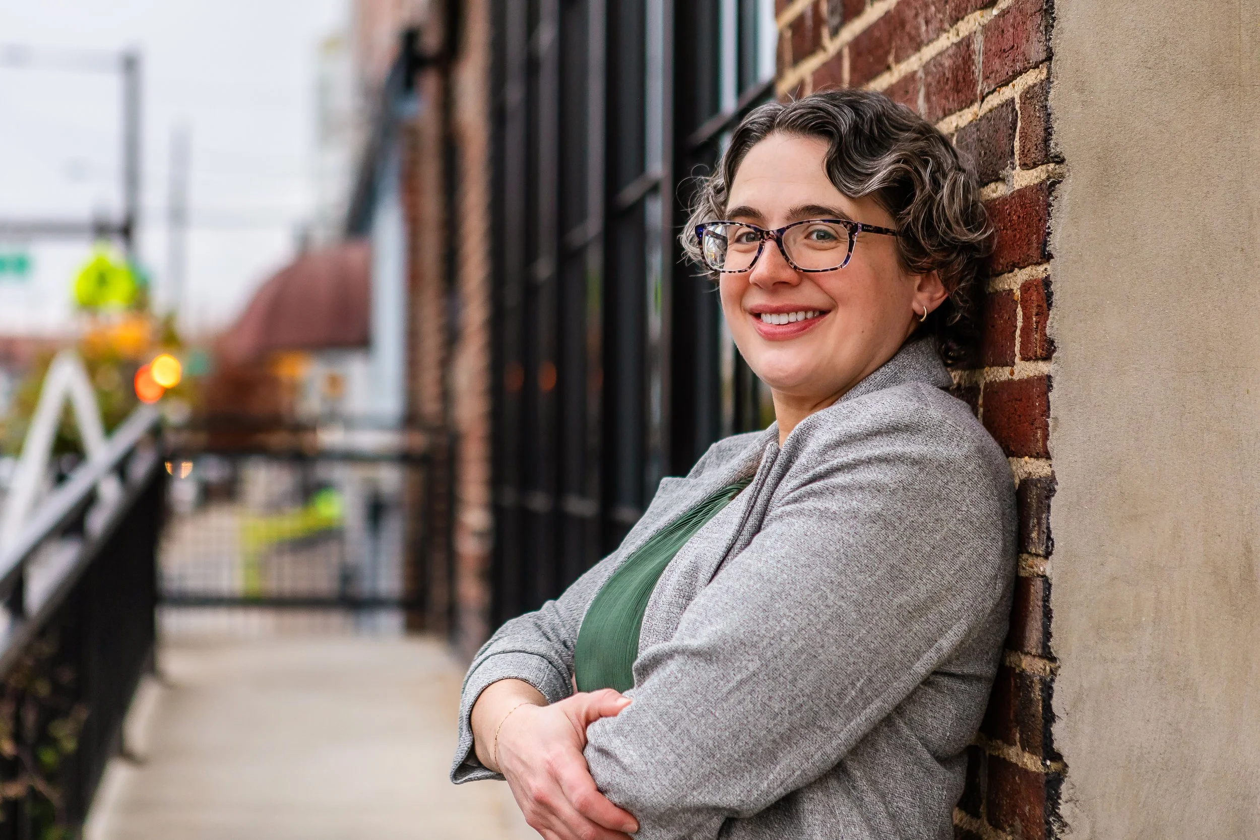 A woman with short curly hair, glasses, and a gray blazer leaning against a brick wall, smiling with her arms crossed.