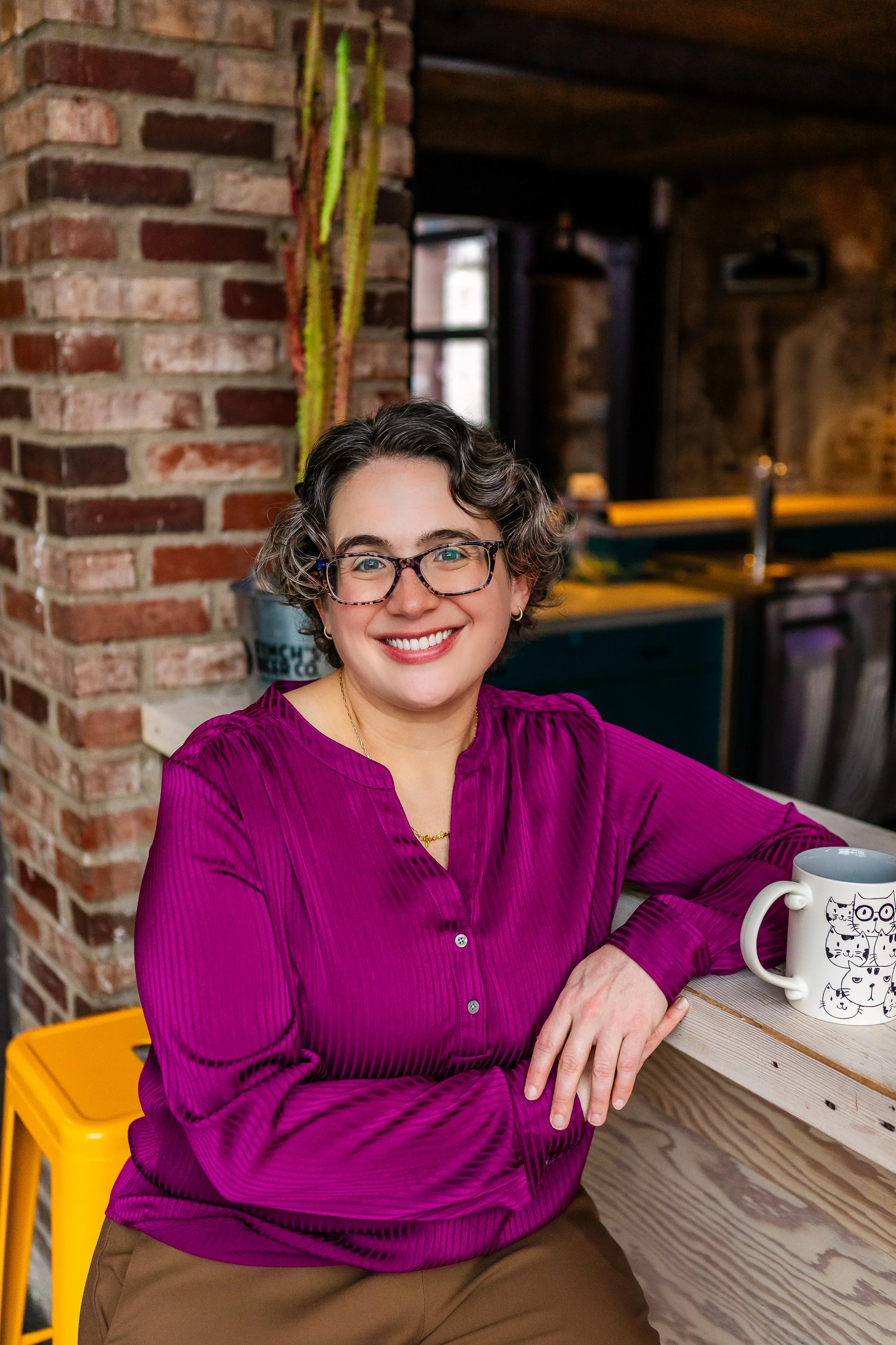 A woman with short curly hair and glasses, smiling, sitting at a wooden table in a cozy, rustic cafe with brick walls and a cactus in the background, wearing a magenta blouse and brown pants.
