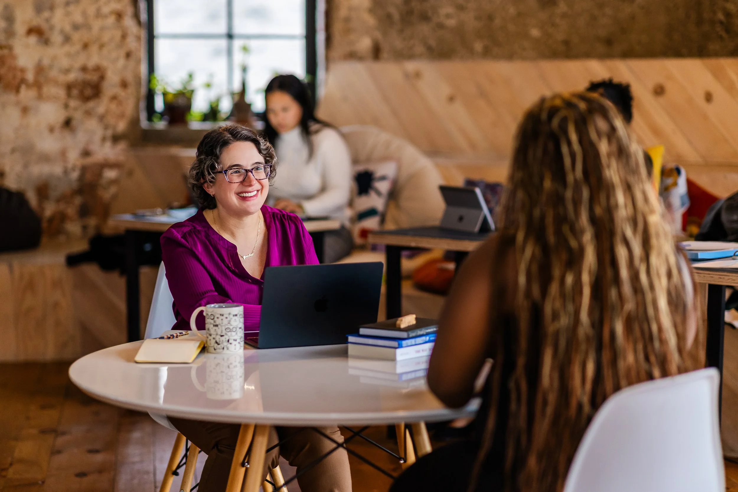 A woman with glasses and short curly hair, sitting at a round table, smiling while talking to another woman with long curly hair. The table has a laptop, a mug, notebooks, and books. In the background, a woman with long dark hair is sitting by a window, working.