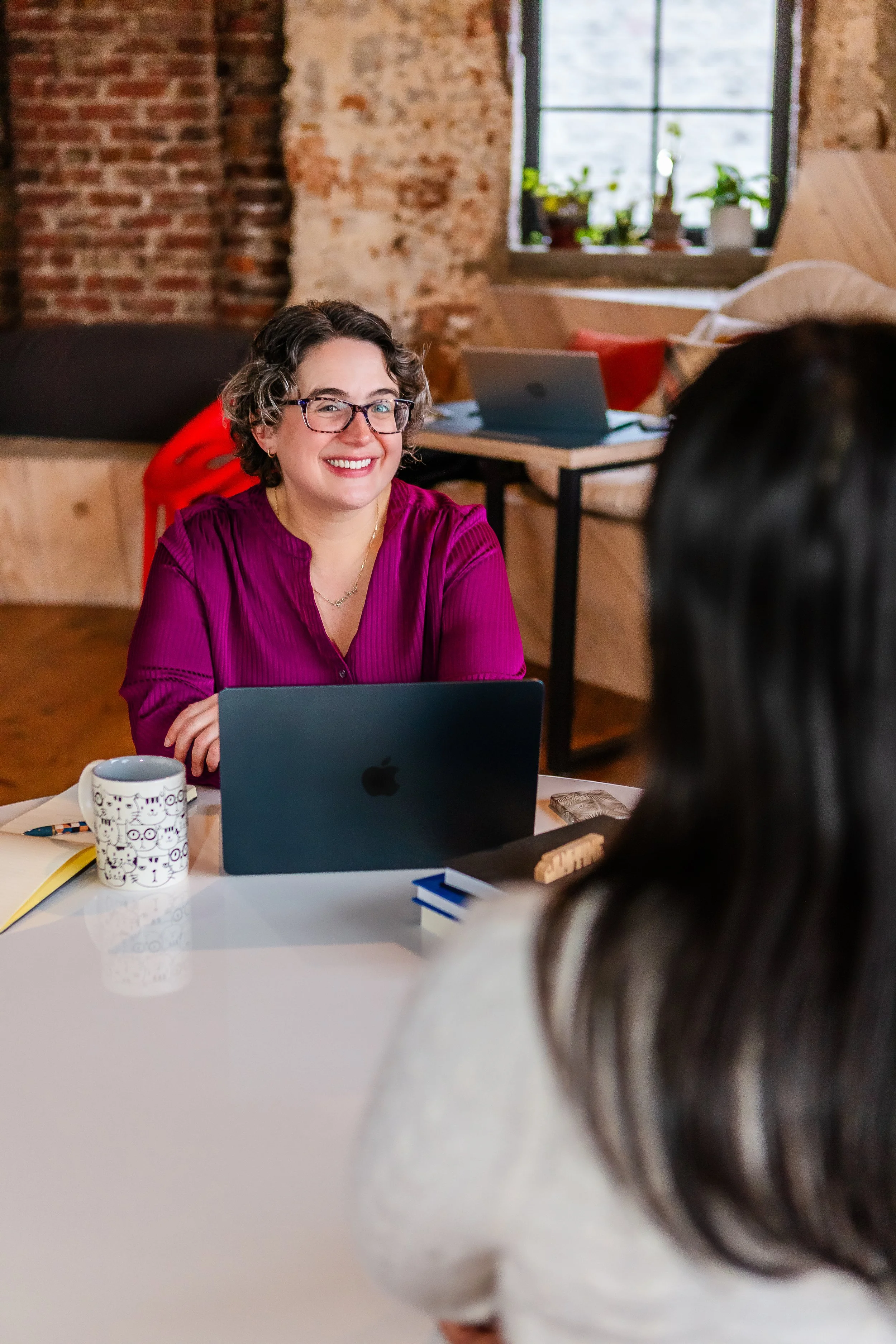 A woman with curly hair, glasses, and a purple blouse sitting at a table with an open laptop, smiling and talking to another person whose back is to the camera. The setting is a cozy, modern space with exposed brick walls and a window with plants.