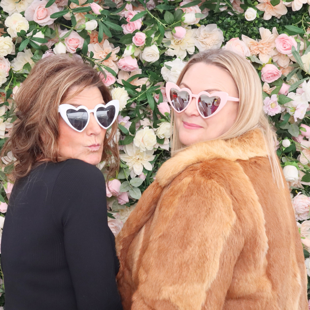 Two women wearing heart-shaped sunglasses posing in front of a floral background. This is in front of a photobooth, provided by Royale Booths photobooth hire in Auckland.