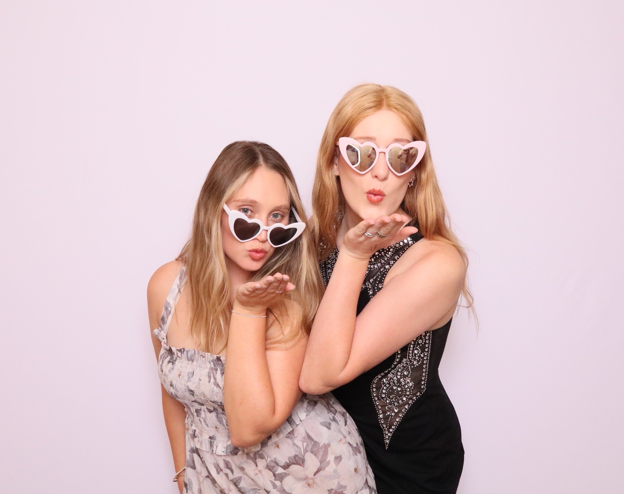 Two women wearing large heart-shaped sunglasses blowing kisses at the camera against a plain white background. This is taken on a professional photobooth by Royale Booths Photobooth Hire.